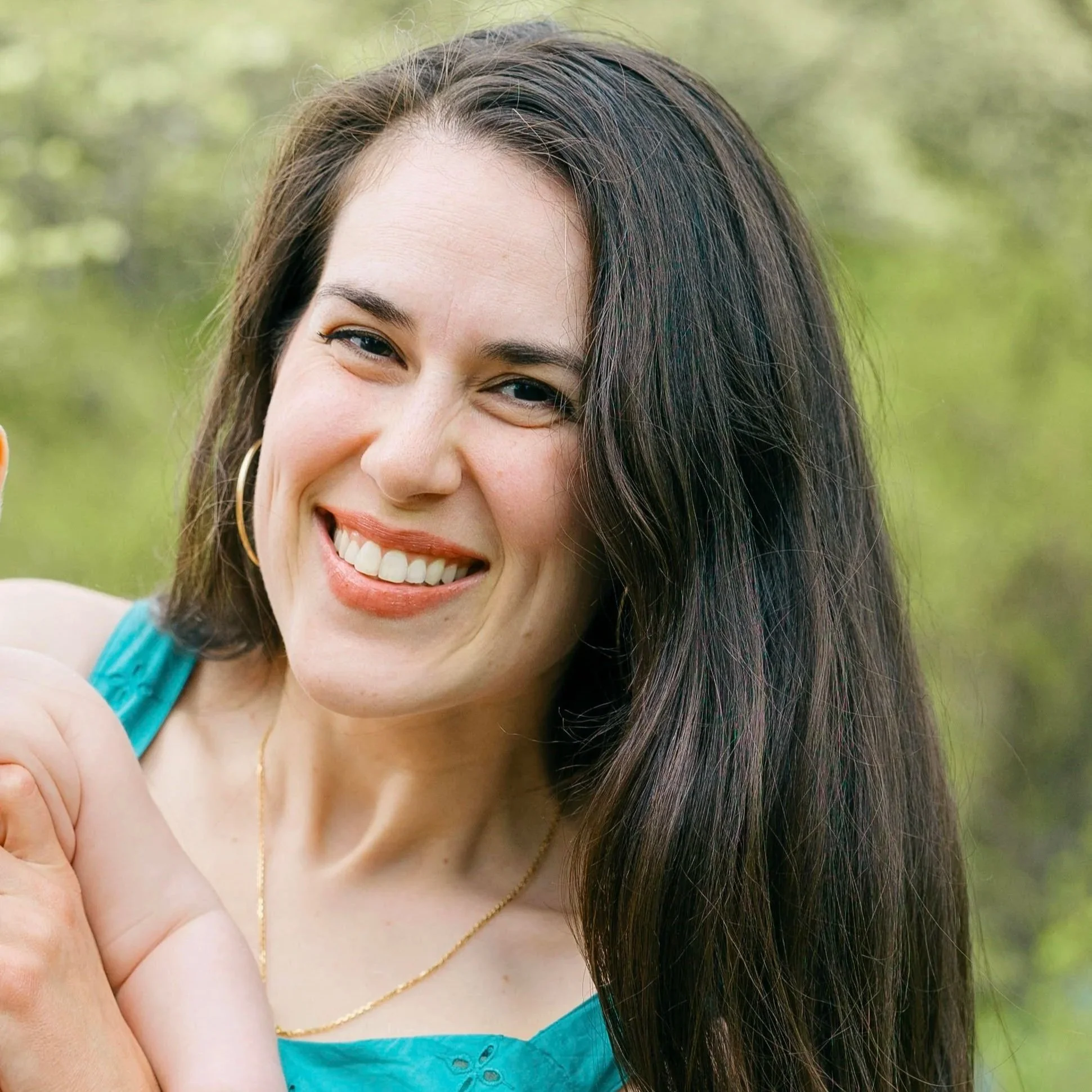 A woman with long dark hair, wearing hoop earrings, smiling outdoors.