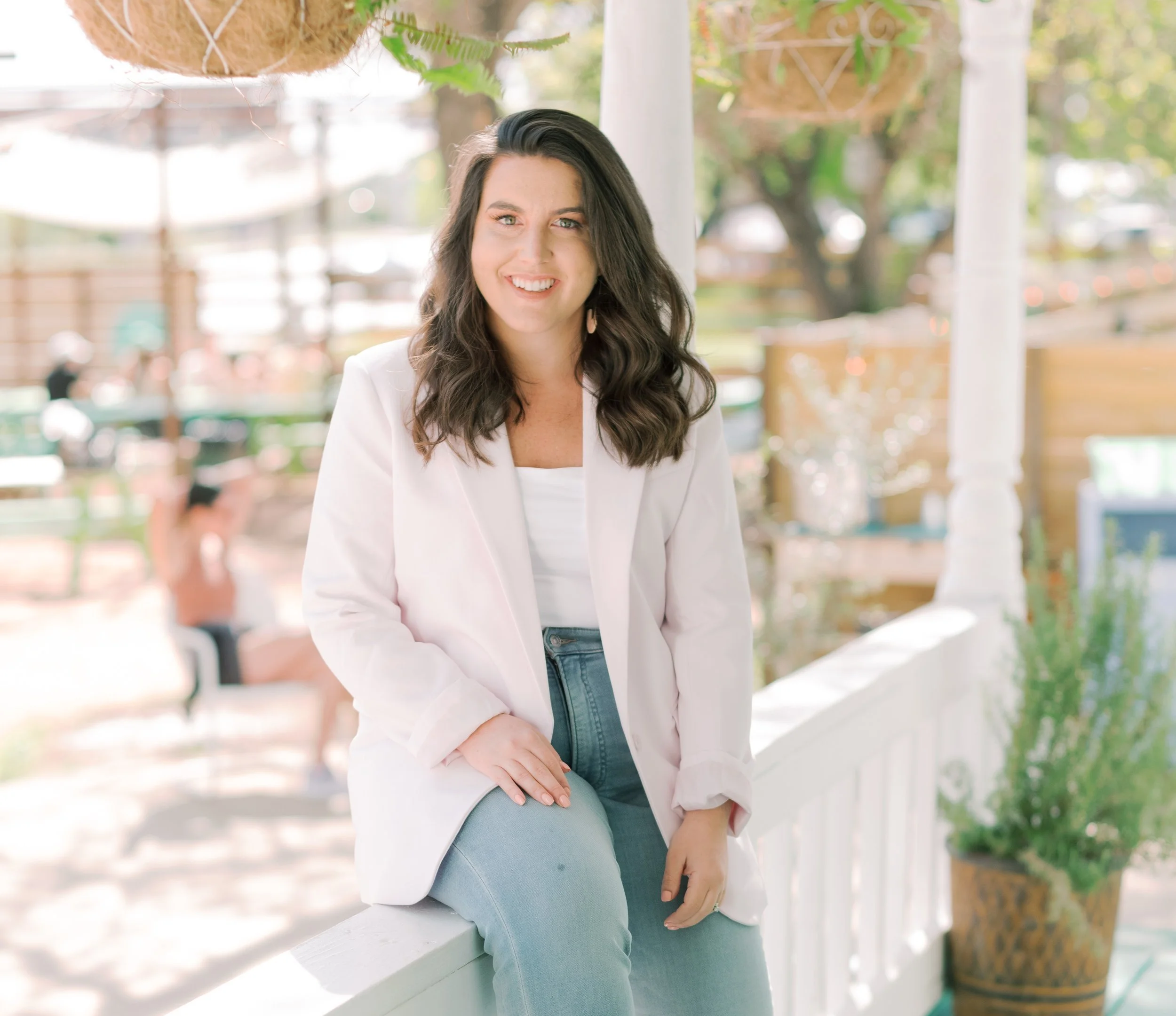 A woman with dark, curly hair smiling while sitting on a white railing outdoors, with a blurred background of people and trees.