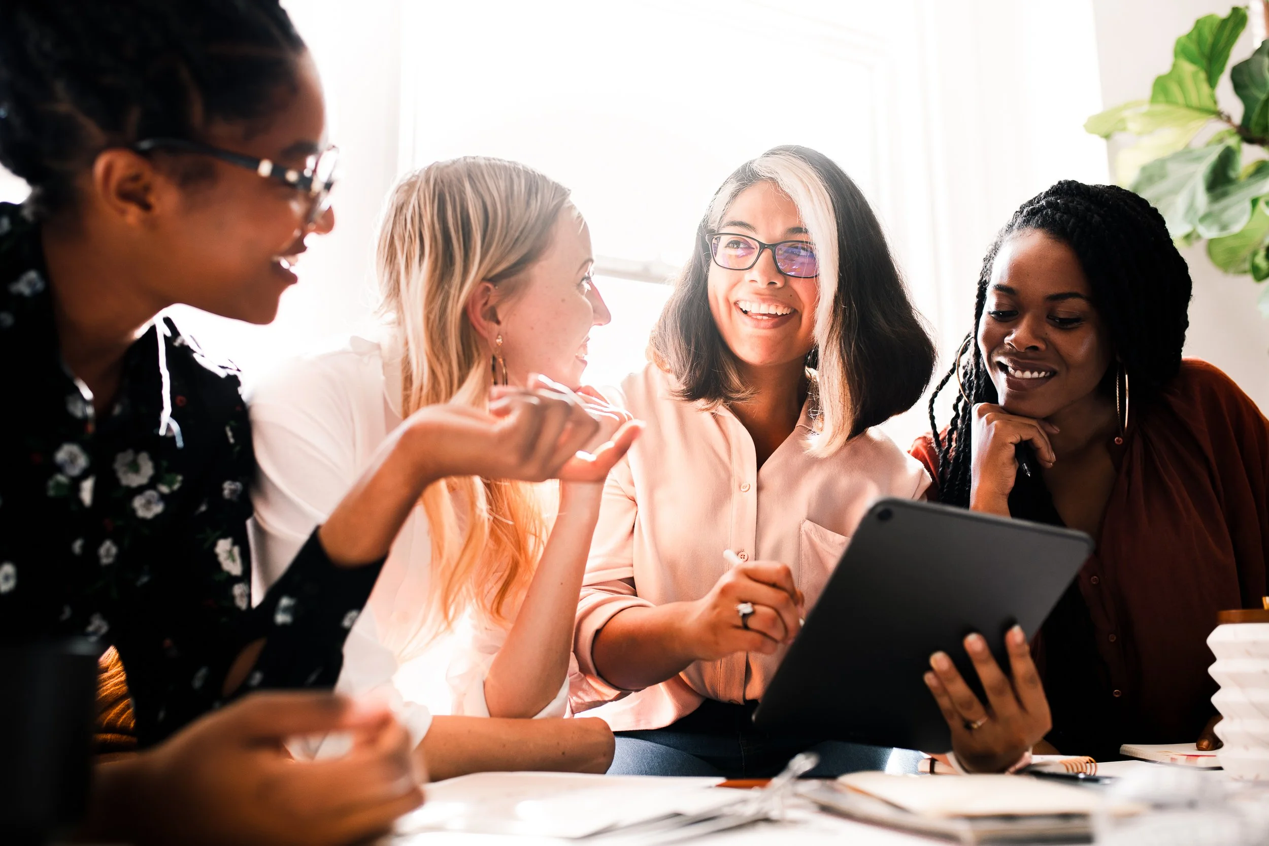 Four women sitting together, looking at a tablet and laughing, in a bright room with a window and plant in the background.