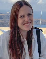 A woman with long brown hair smiling outdoors near a body of water with mountains in the background.