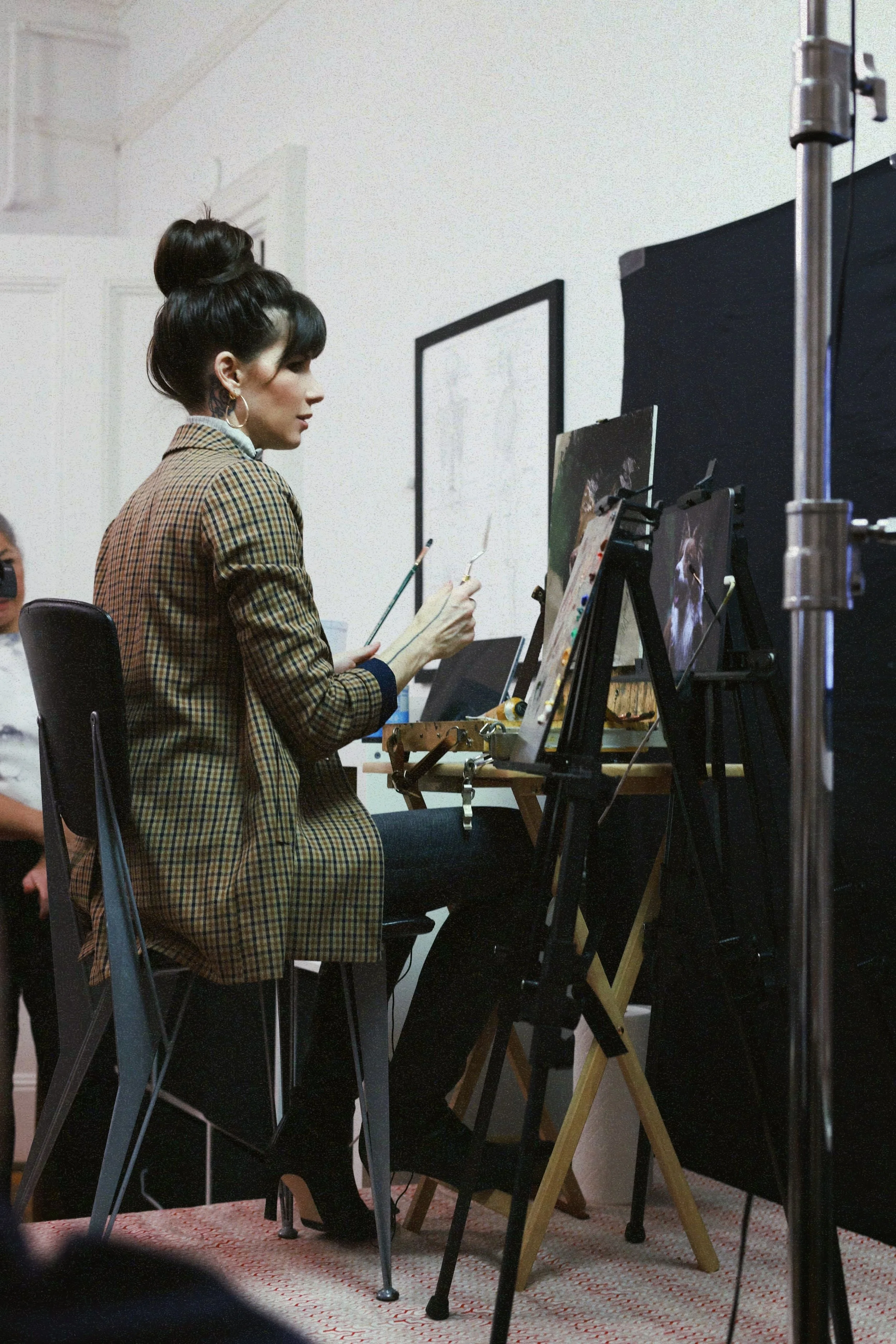 A woman with dark hair styled in a high bun, wearing a brown plaid blazer and hoop earrings, is seated in front of an easel with a painting. She is holding a palette and paintbrushes, and she appears to be working in an art studio or classroom with frames and equipment visible on the wall.