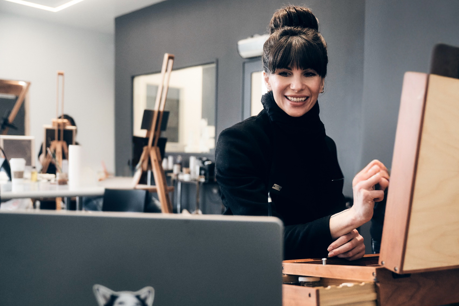 A woman with dark hair in a bun, smiling, wearing a black jacket, sitting at an easel in an art studio with multiple easels and art supplies in the background.