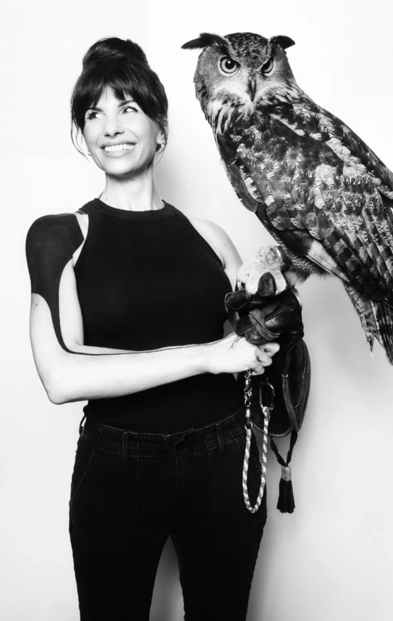 A woman with short dark hair, wearing a sleeveless black top, is holding a large owl on her arm. The owl has intricate feather patterns and is wearing a glove on its talon. The image is in black and white.