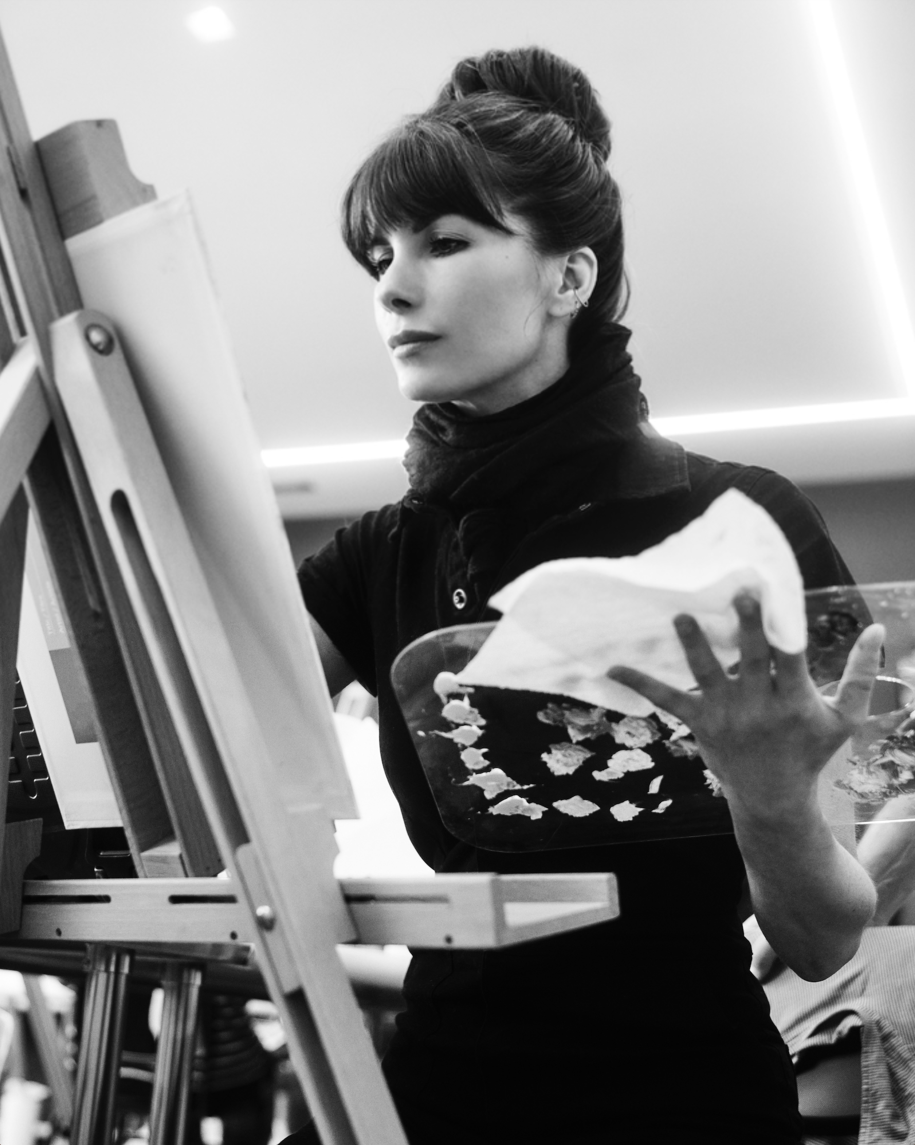 A woman with dark hair styled up, wearing a dark top with light-colored patches on the sleeves, is painting on a canvas in an art studio.