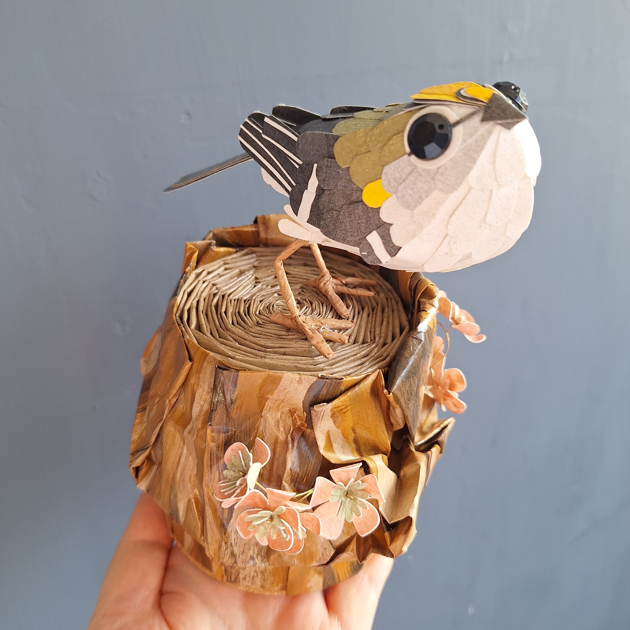 Handmade paper sculpture of a goldcrest bird perched on a tree stump. Original paper art by Kaper