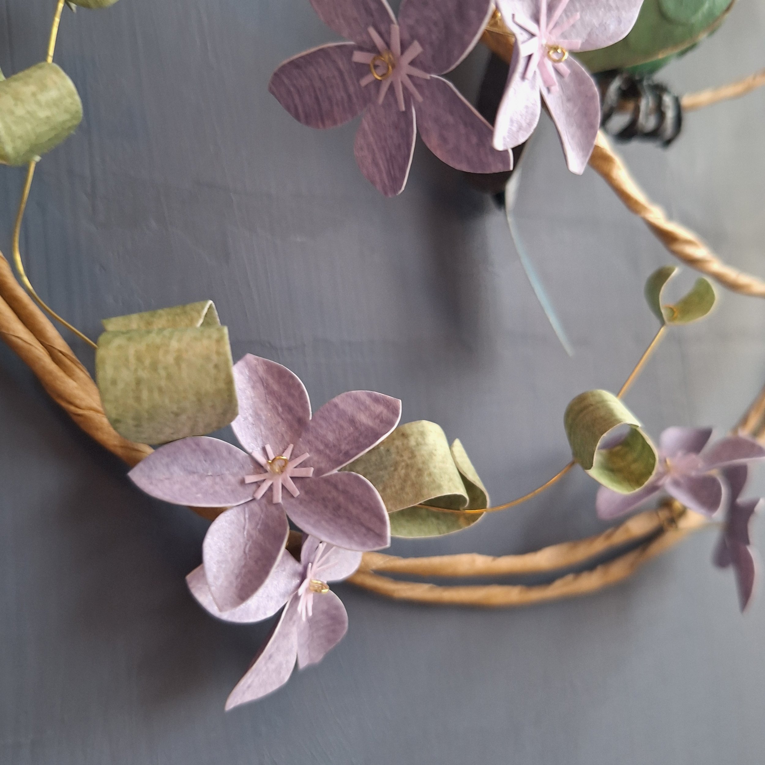 This violetear hummingbird sculpture perches on a wreath of purple flowers and pale green leafy stems. This paper sculpture is one of a kind.
