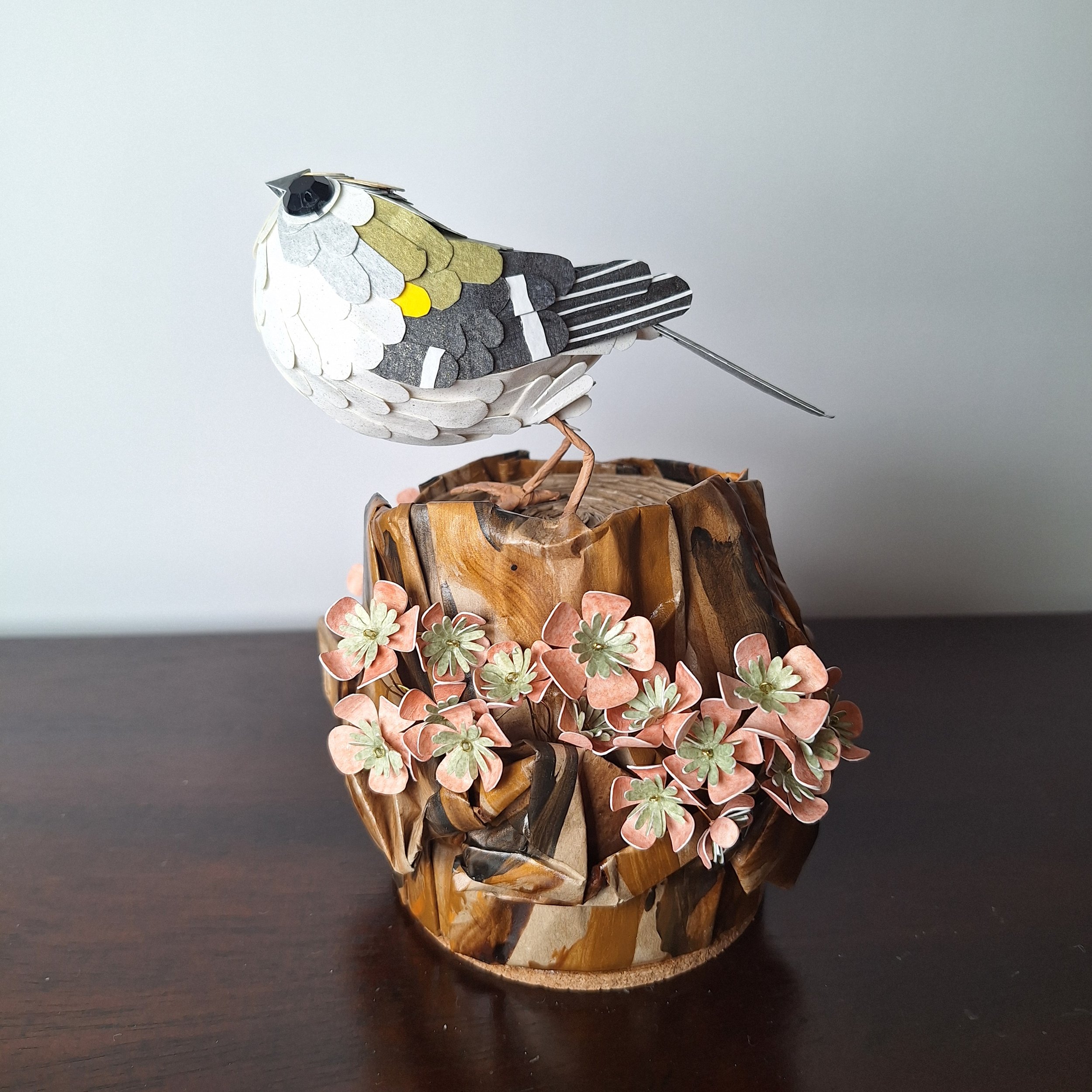 a paper sculpture of a goldcrest bird perches on a handmade tree stump sculpture