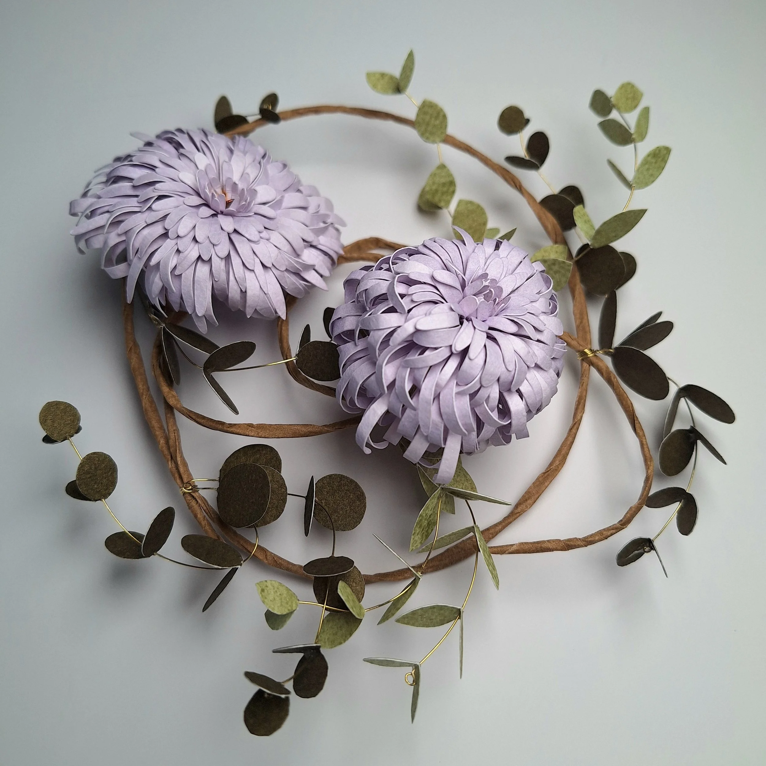 A handmade paper flower wreath, depicting two pale purple chrysanthemums surrounded by green leafy foliage.