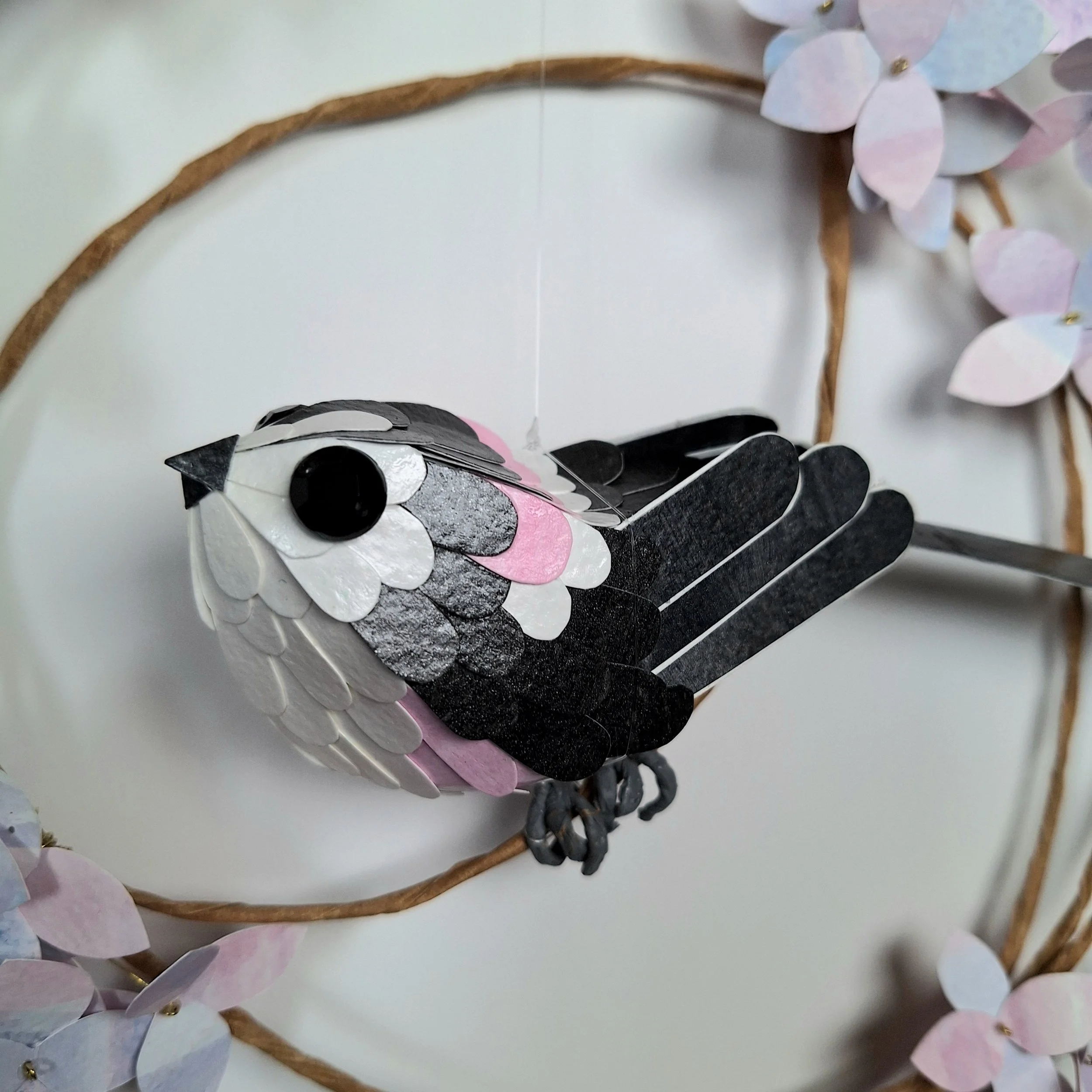close up of the paper bird sculpture, depicting a long tailed tit. He is mostly black and white, with a tuft of pink at the top of his neck and under his wings.