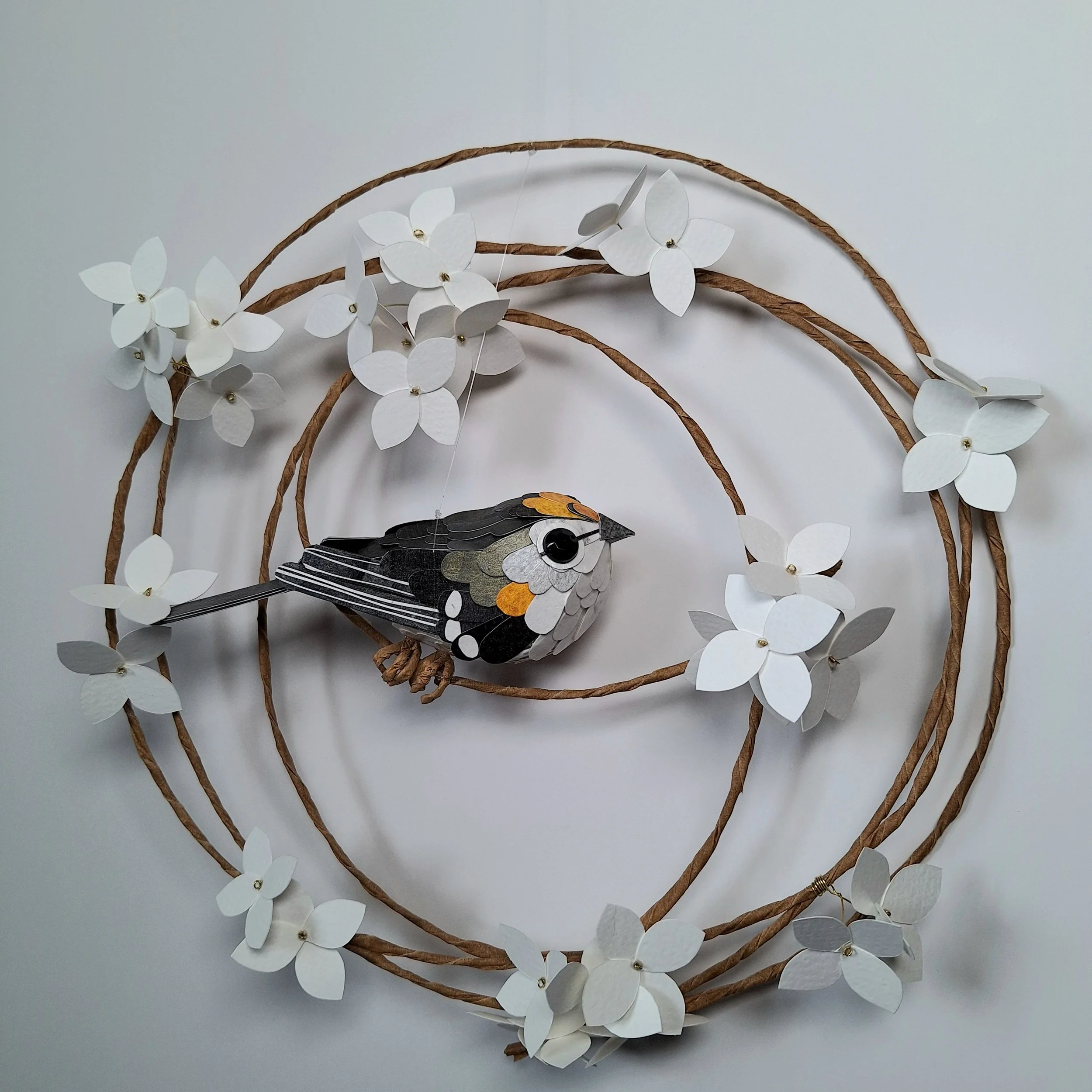 A handmade paper sculpture of a floral wreath with a tiny goldcrest bird perched in the middle of the wreath, surrounded by crisp white hydrangea blooms.