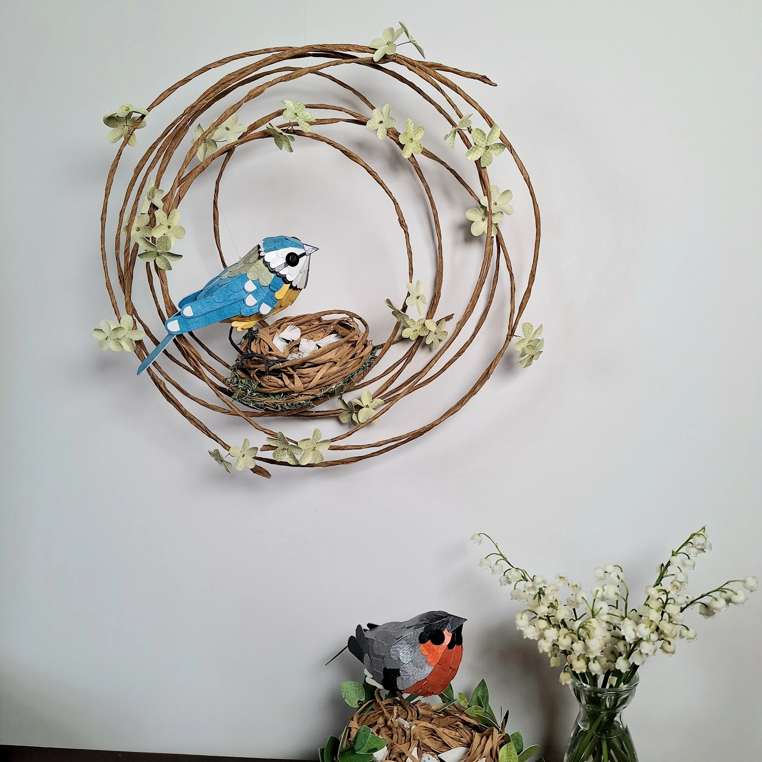 A bird sculpture featuring a blue tit perched on a nest on a green floral wreath hangs on a wall.