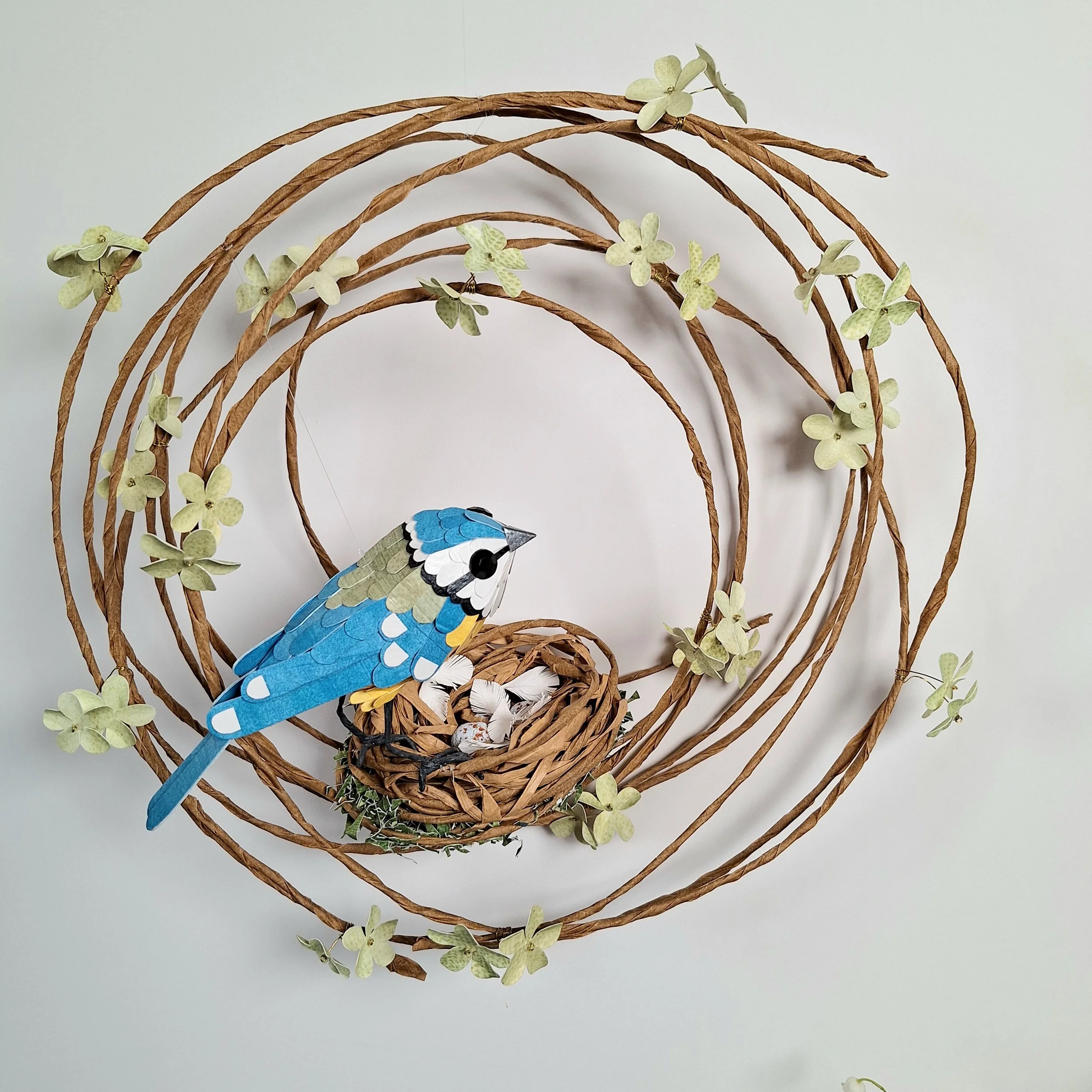 A blue tit perches on a nest adorned with lichen. The nest sits on a floral wreath. This handmade sculpture is made from paper.