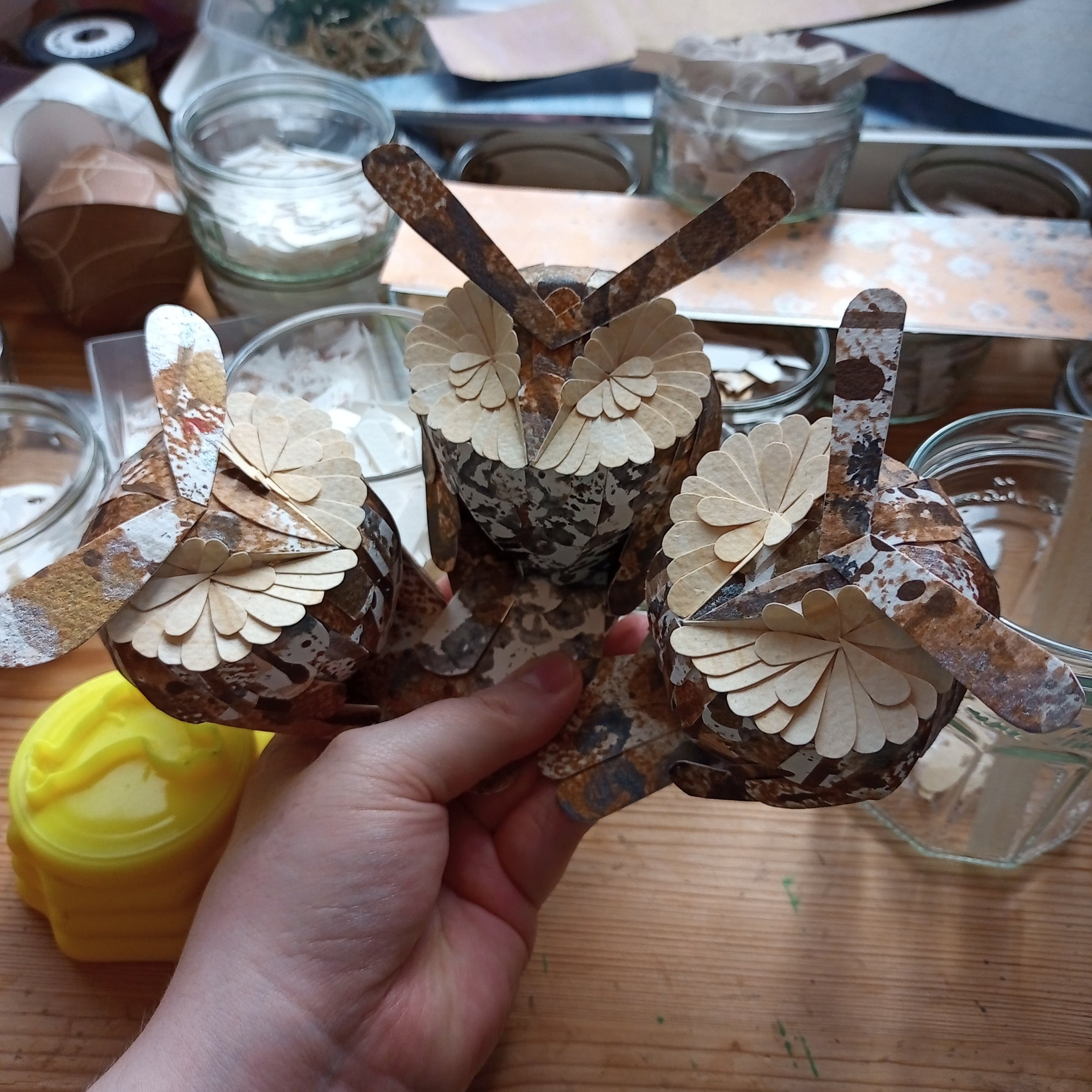 Three handmade paper owls with textured feathers, held in a person's hand, surrounded by craft supplies in glass jars on a wooden table.