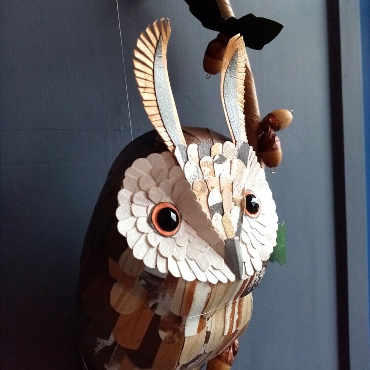 Decorative owl sculpture made of layered wood pieces with large eyes and prominent ears against a dark background.