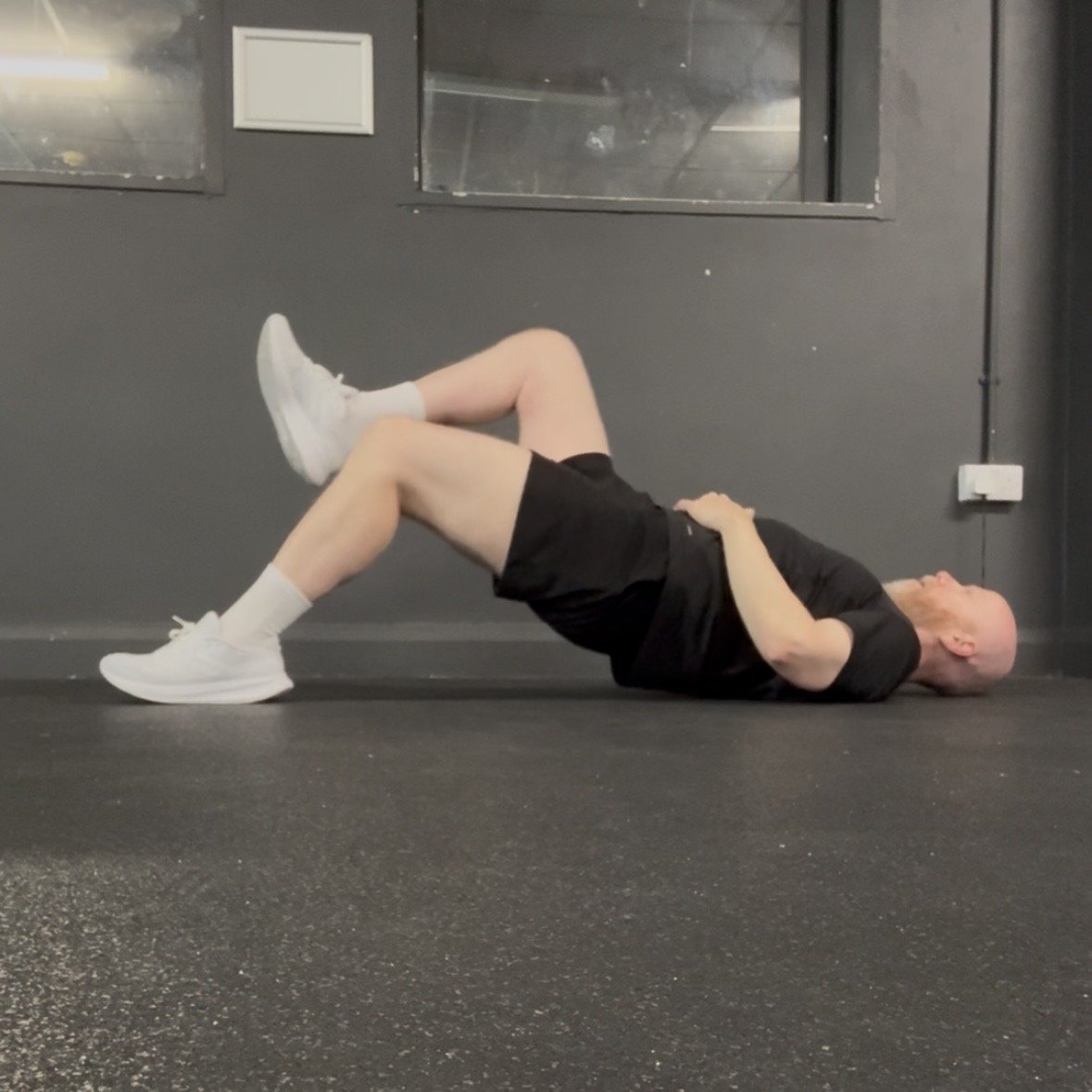 Picture of Andy doing a single leg bridge, he wears a black t-shirt, black shorts and white trainers.