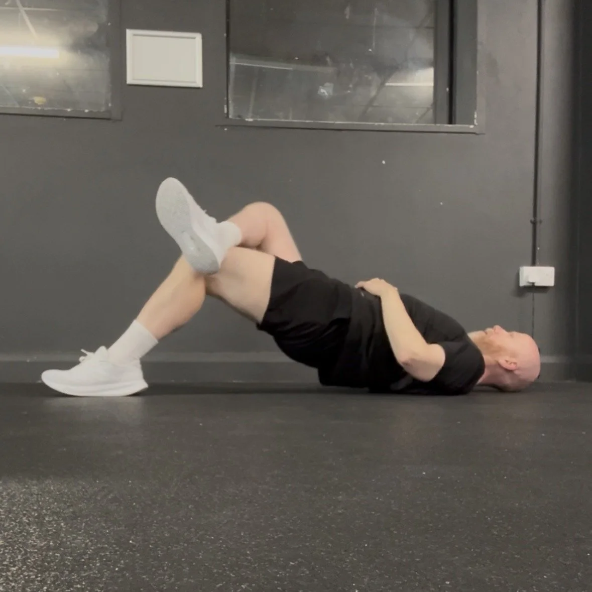 Picture of Andy doing a leg crossed bridge, he wears a black t-shirt, black shorts and white trainers.