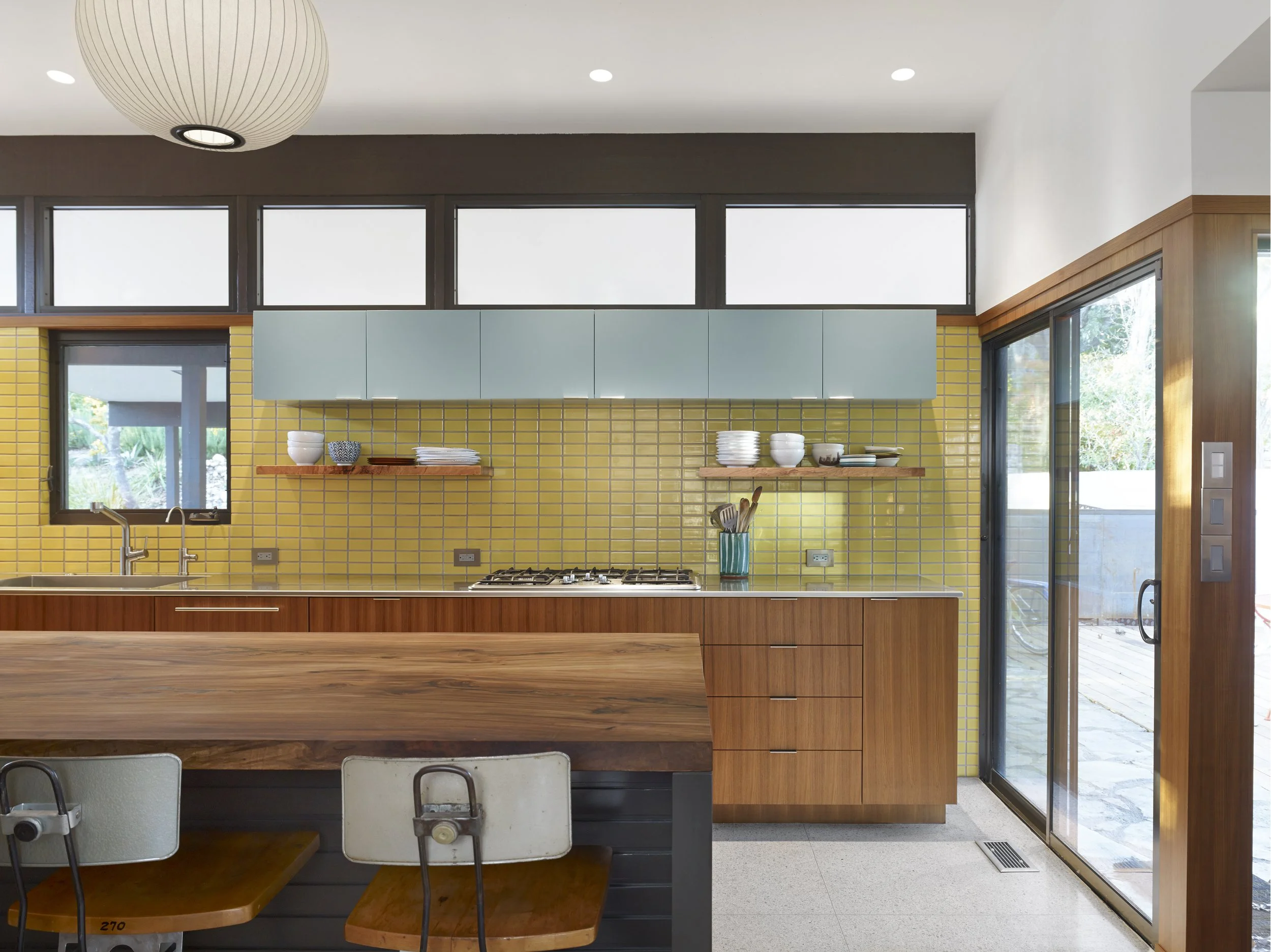 Modern kitchen with yellow tile backsplash, wooden cabinets, blue wall-mounted cabinets, and a sliding glass door leading outside.