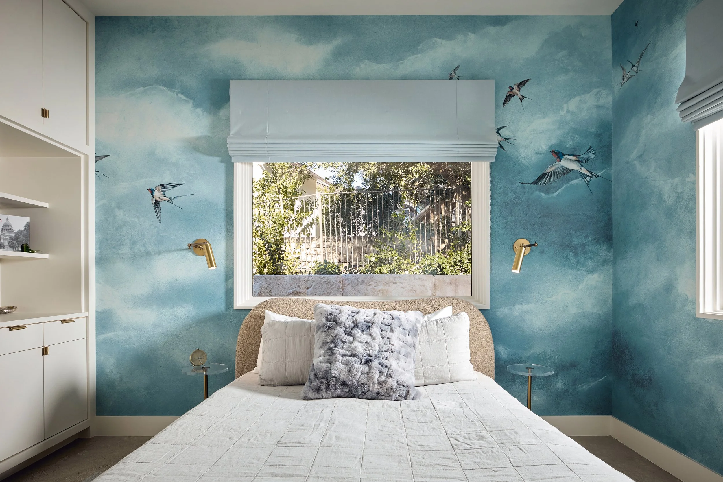 Bedroom with blue sky-themed wall mural of clouds and flying birds, beige headboard, white bedding and pillows, gold wall-mounted reading lights, large window with white blinds, and greenery outside.