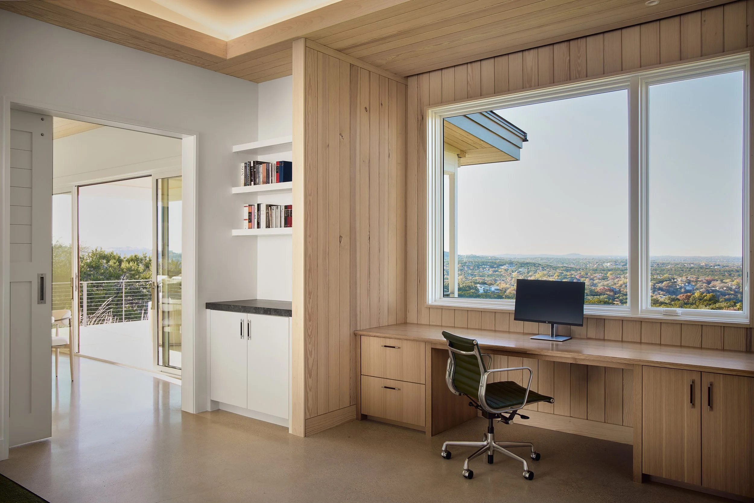 Home office with large window overlooking city view, wooden desk, black office chair, built-in shelves with books, sliding glass door leading to outdoor balcony.
