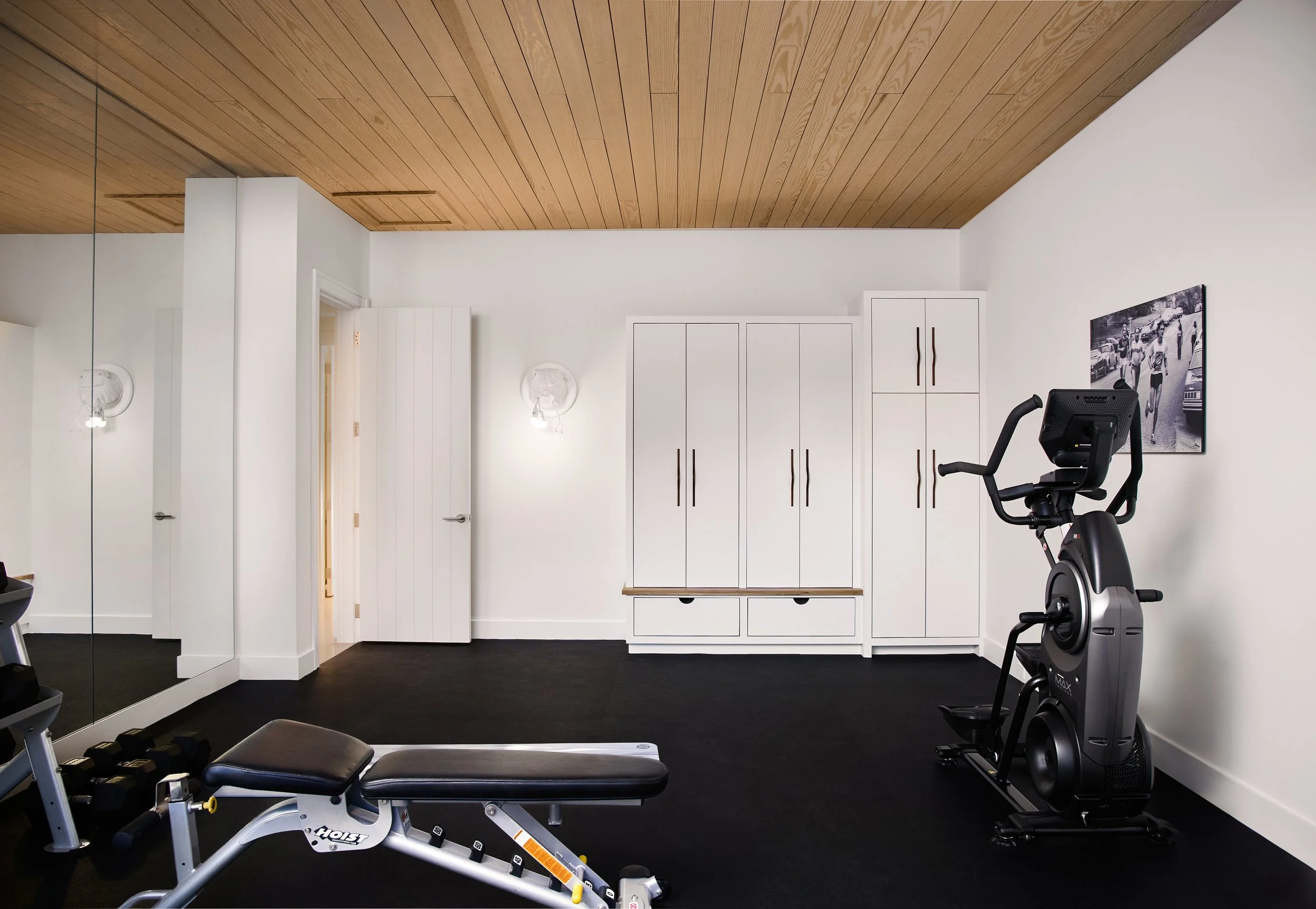 Home gym with black exercise bike, weight bench, and white cabinets, wood-paneled ceiling, mirror, and black floor.