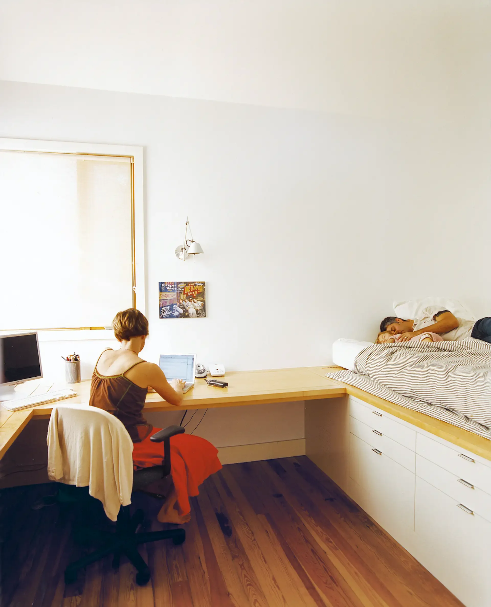 A woman sitting at a desk working on a laptop in a room with a bed on the right side. A man is sleeping in the bed, and there is a window on the left wall with a small wall art piece below a wall lamp.