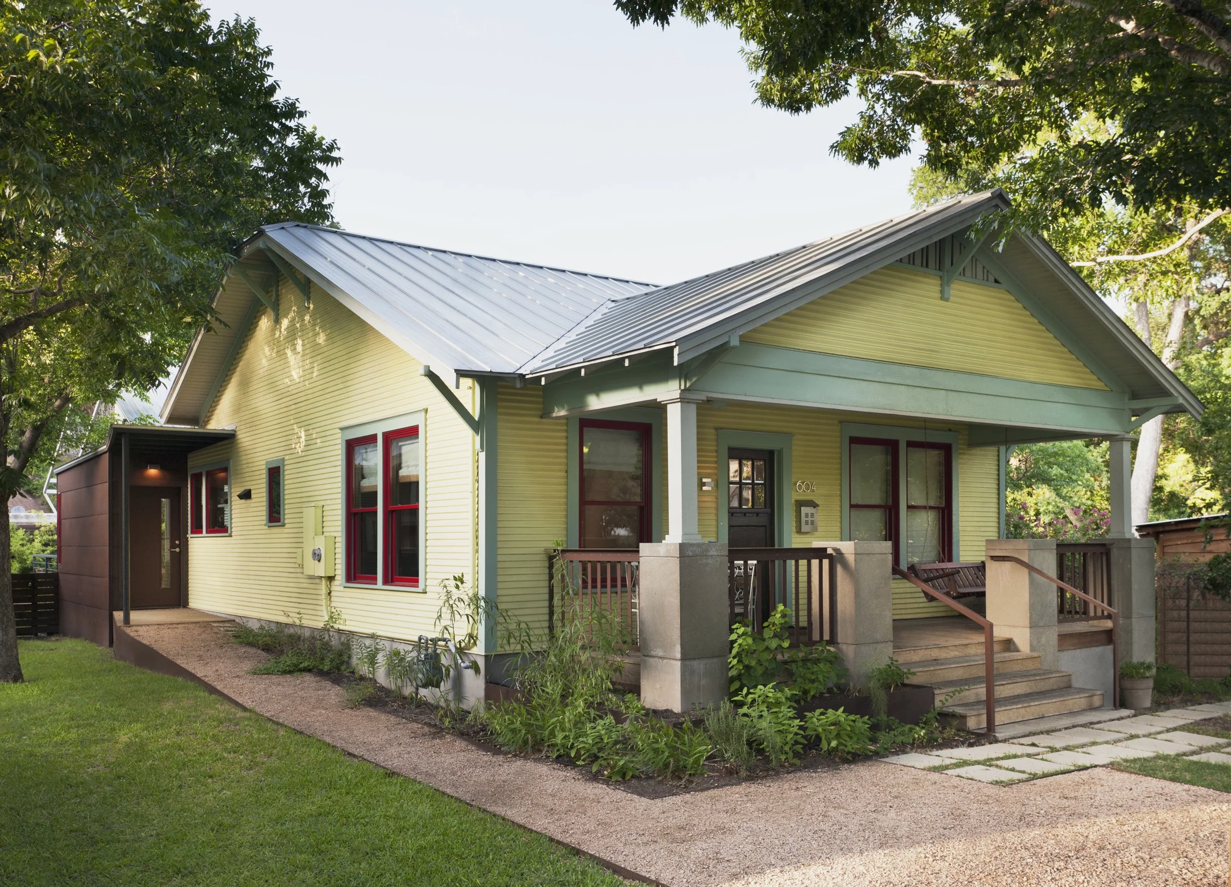 Austin architecture. Classic bungalow, yellow house with a covered porch, gray stairs, green trim, and red window frames, surrounded by greenery.