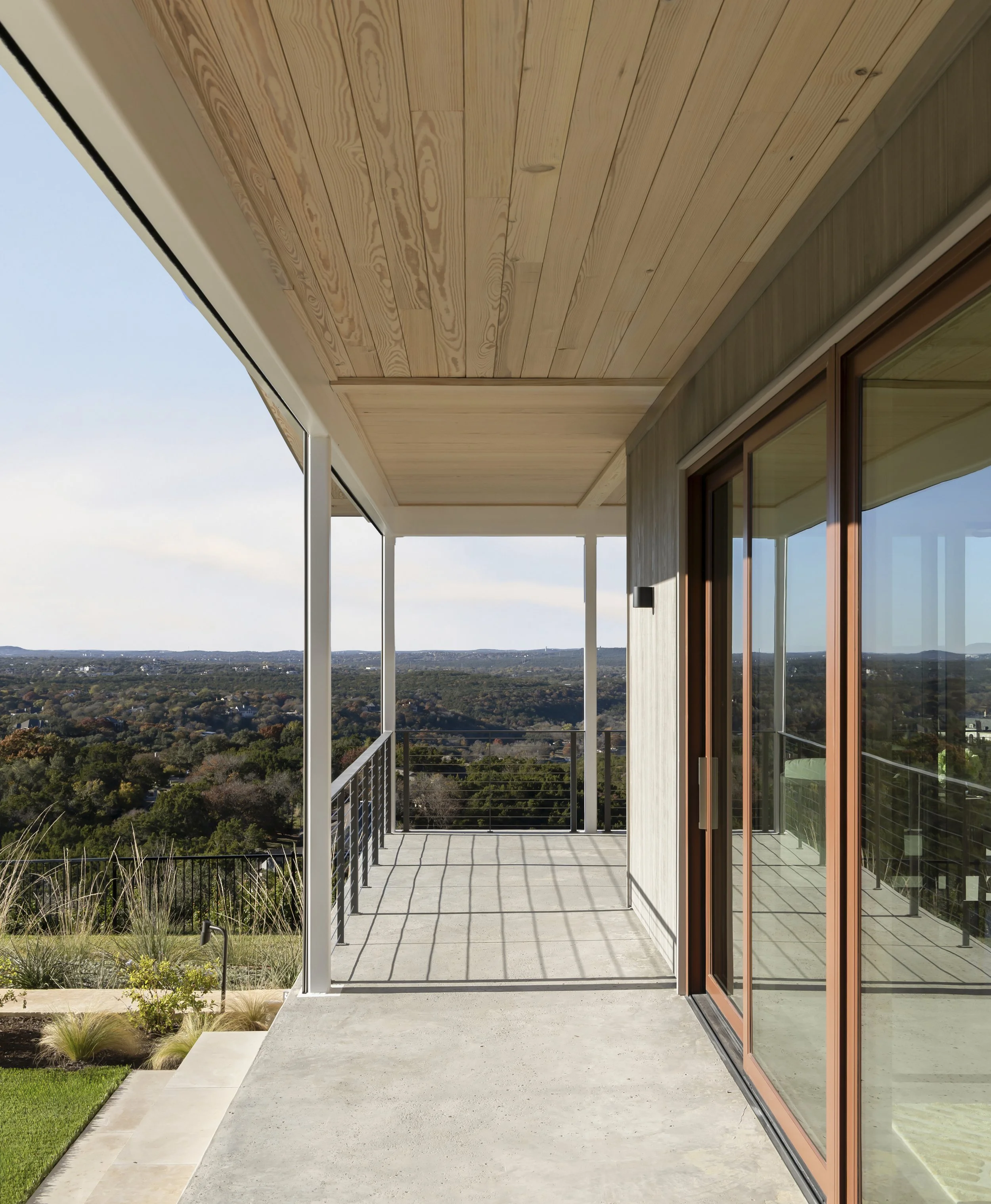 Modern balcony with concrete floor, railing, and sliding glass doors, overlooking a scenic landscape with trees and hills under a partly cloudy sky.