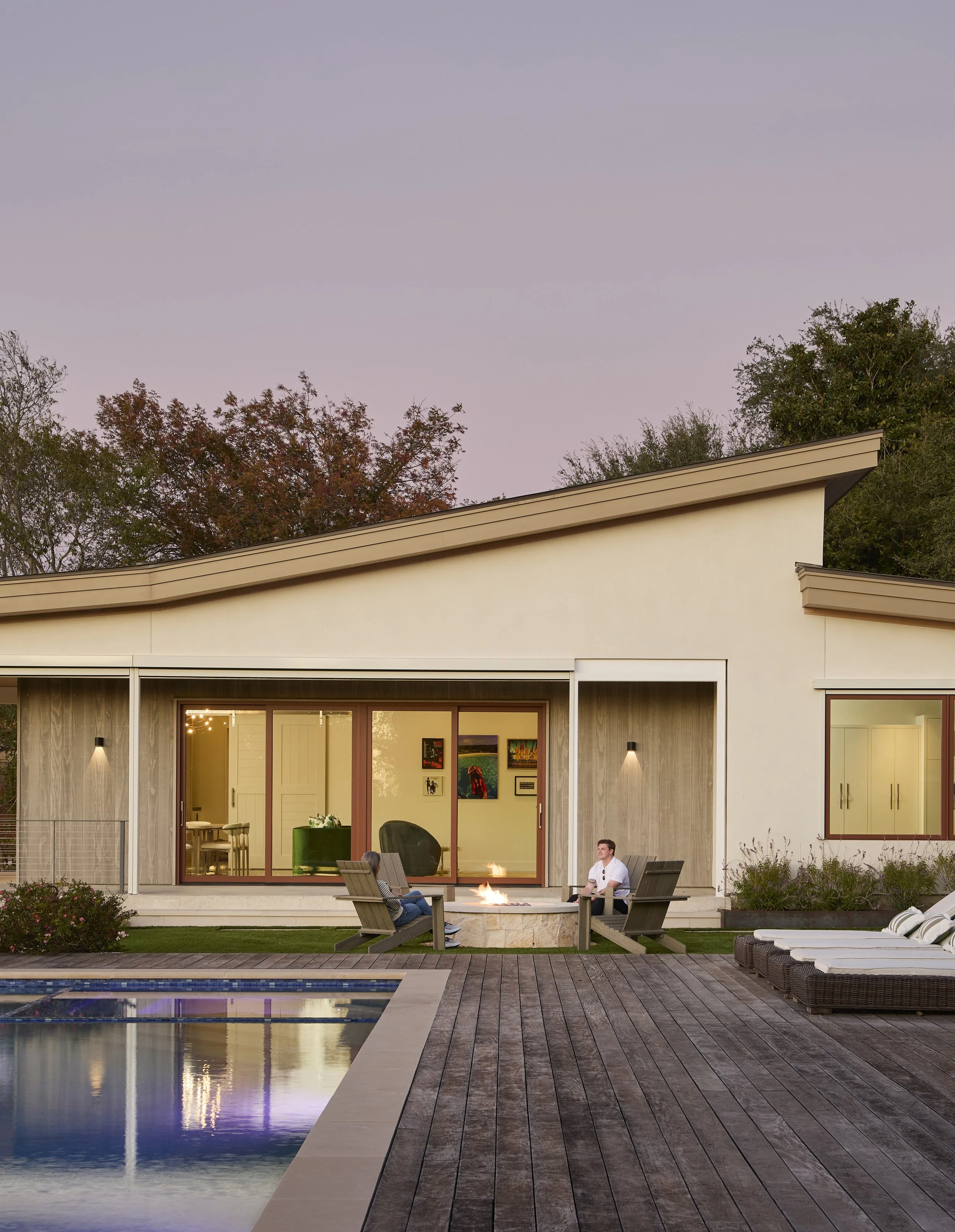 A backyard with a swimming pool, wooden deck, and a modern single-story house with large glass doors. Two people are sitting by a fire pit, talking. There are patio chairs, lounge chairs, and some plants around.
