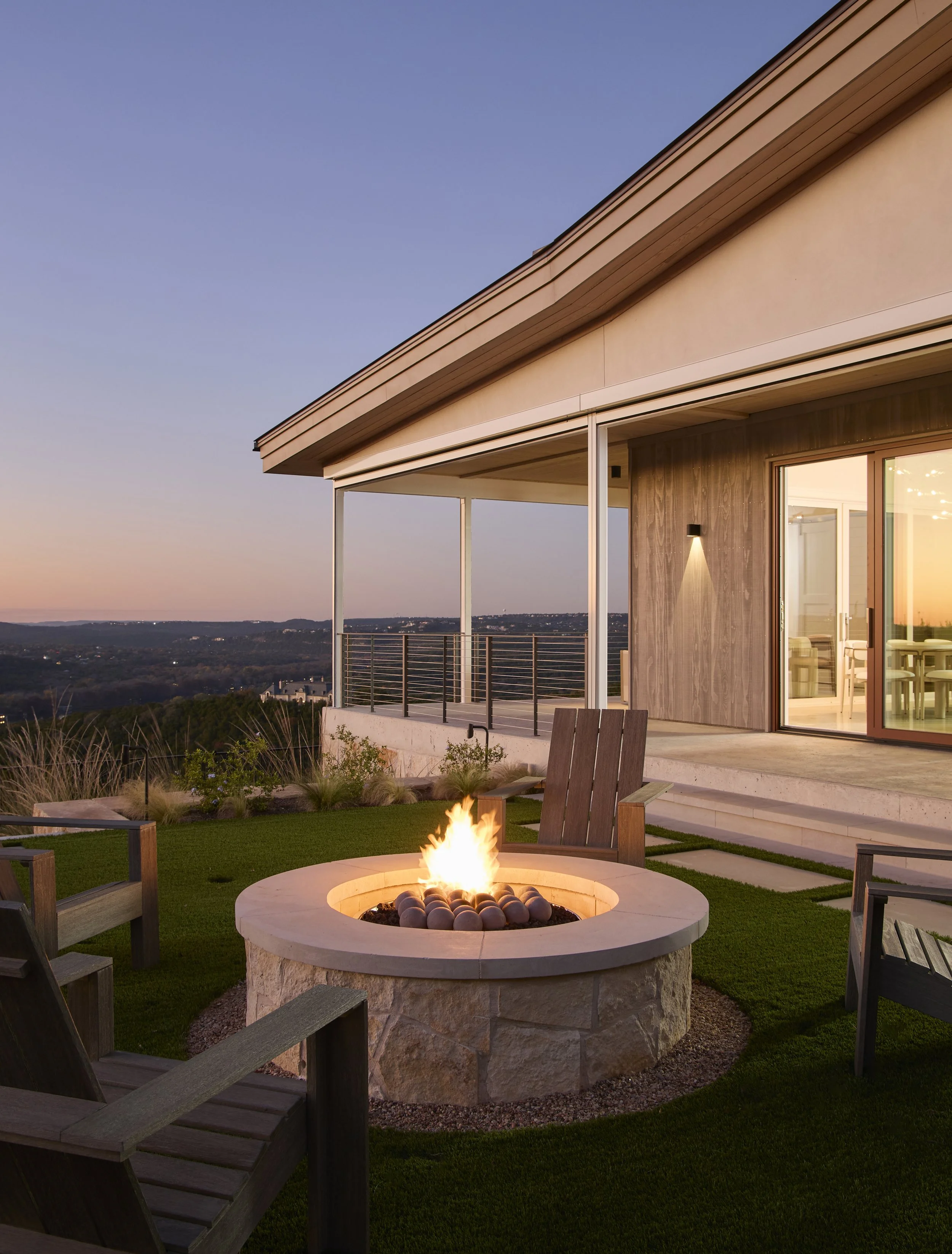 Outdoor patio area with a fire pit, wooden chairs, grass, and a modern house with large glass doors at sunset.
