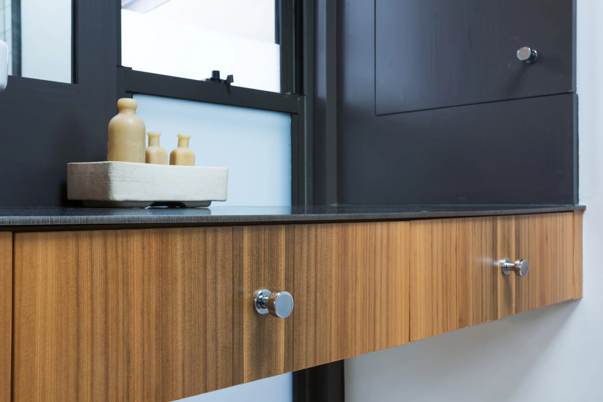 A wooden cabinet with two drawers and round metal handles, black countertop, a white tray with three beige ceramic bottles, and a window with black frame in the background.