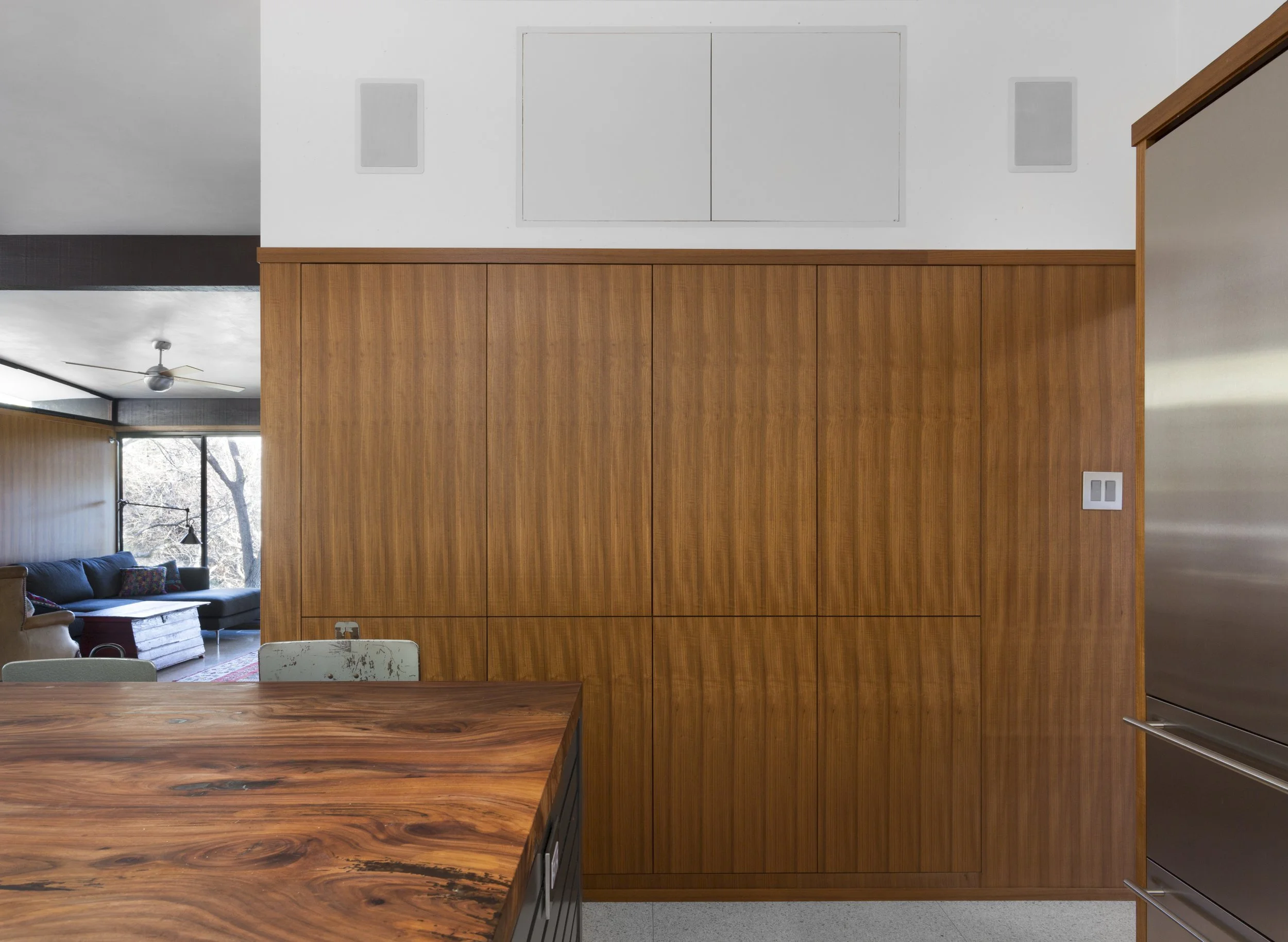 Interior view of a modern kitchen with wooden cabinetry, a partial view of a dining table in front, and a glimpse of a living room with a sofa and large window with trees outside.