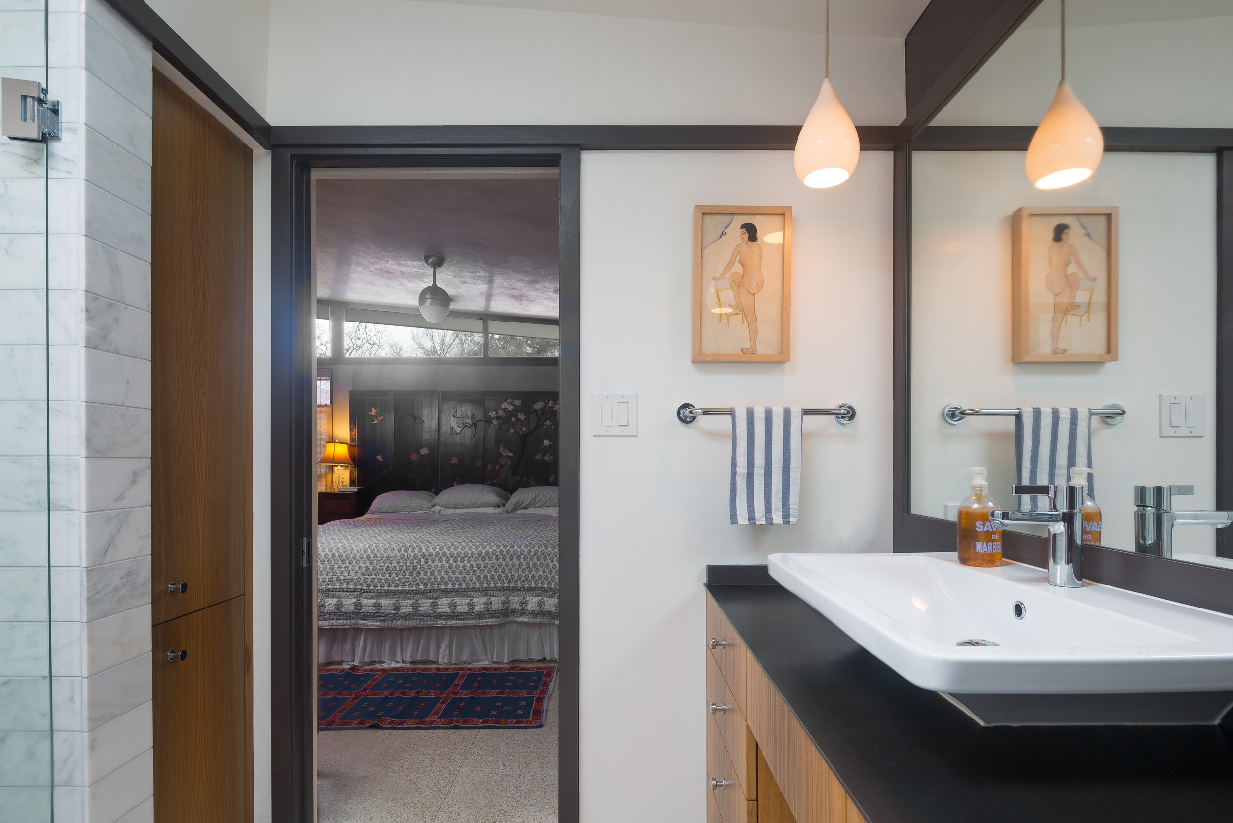 Bathroom with a large mirror, sink with soap dispenser, wall art, and a view into a bedroom with a bed, nightstand, lamps, and window.