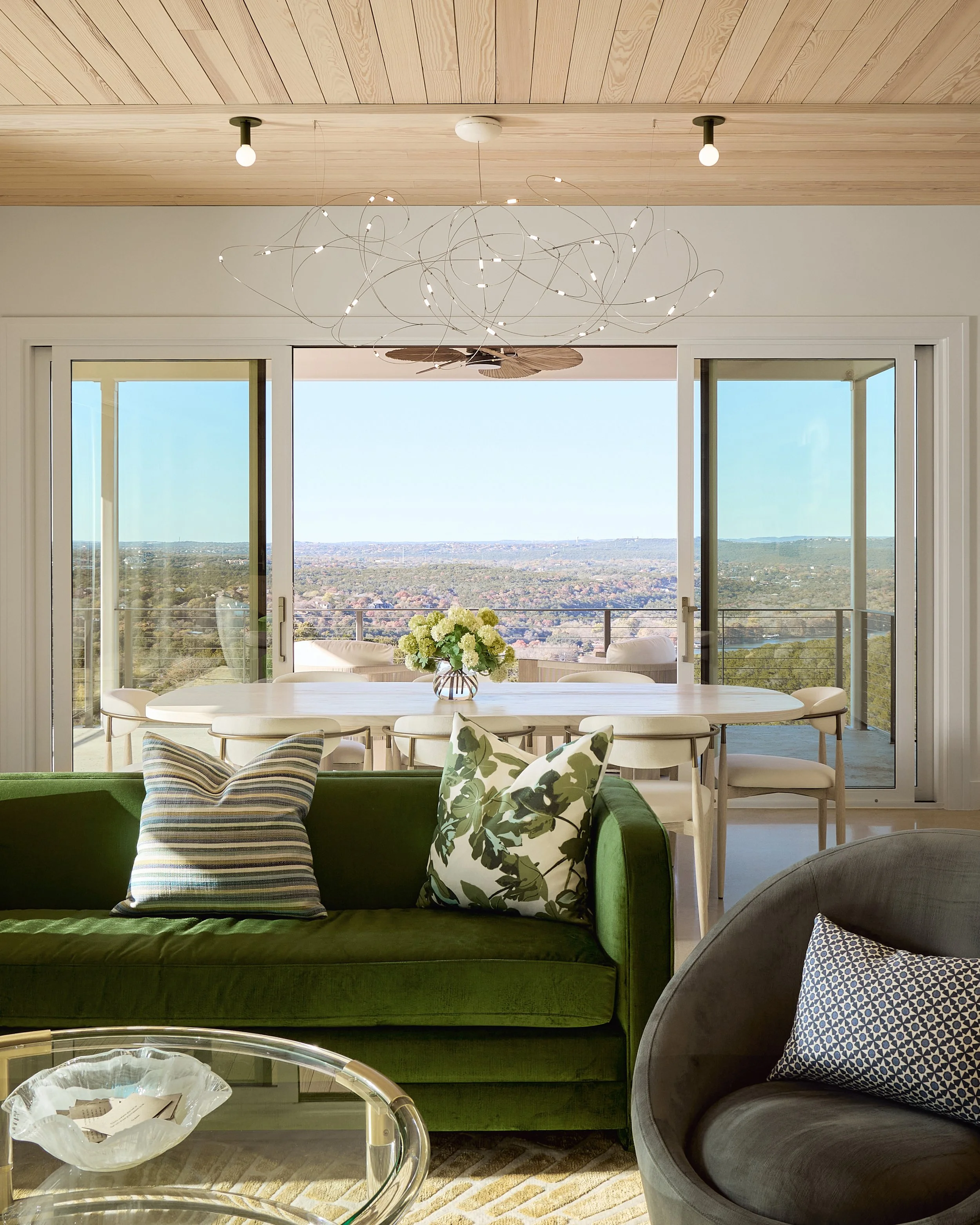 Living room with a green velvet sofa and a gray rounded armchair, decorative pillows, glass coffee table, sliding glass doors opening to a balcony with view of a cityscape and distant hills, ceiling light fixtures, and a modern chandelier.