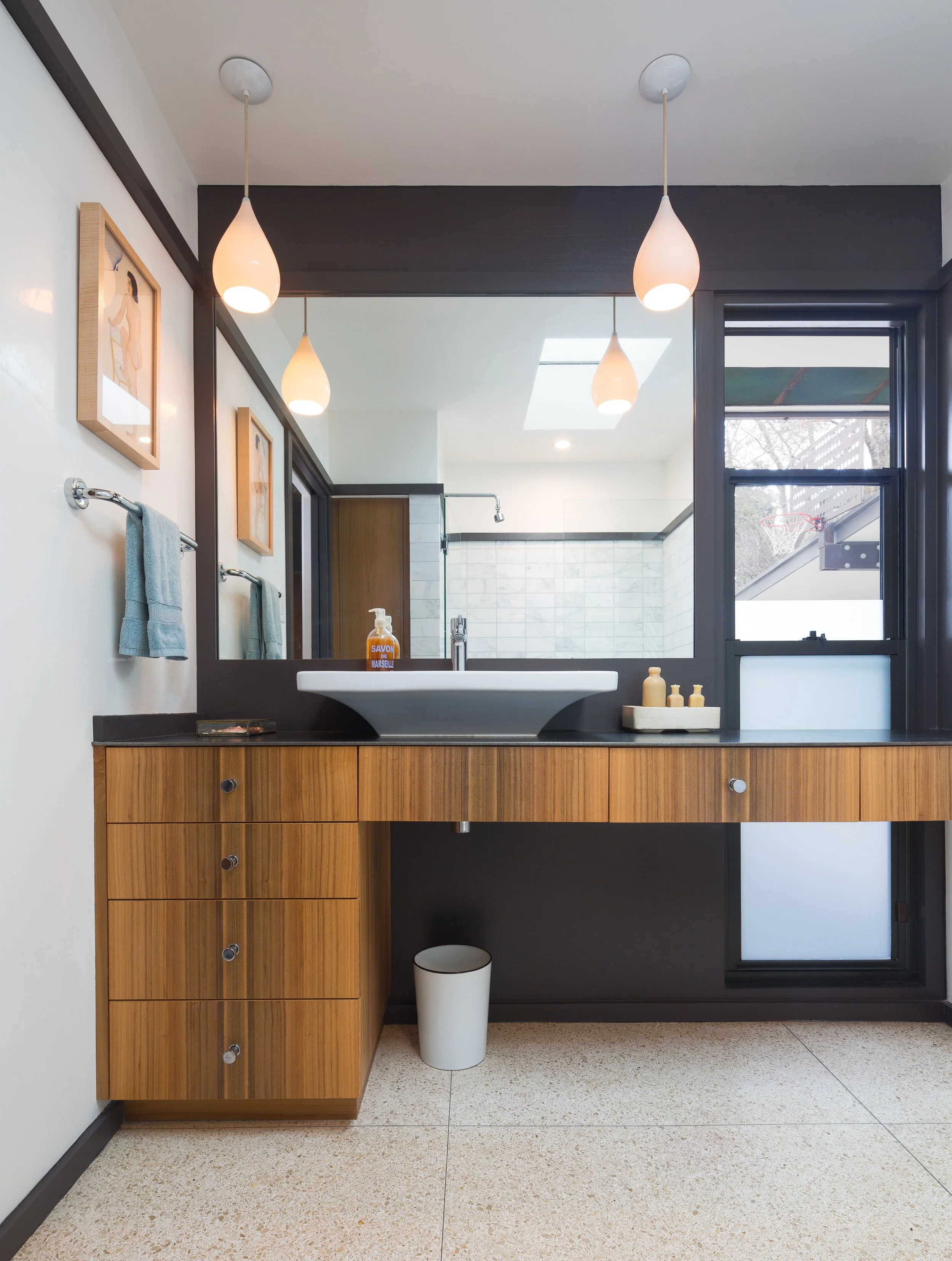 Modern bathroom with a wooden vanity, a white sink, and a large mirror. Three pendant lights hang from the ceiling. There are two teal towels on towel bars and small decorative bottles on the counter. A window and shower are visible in the background.