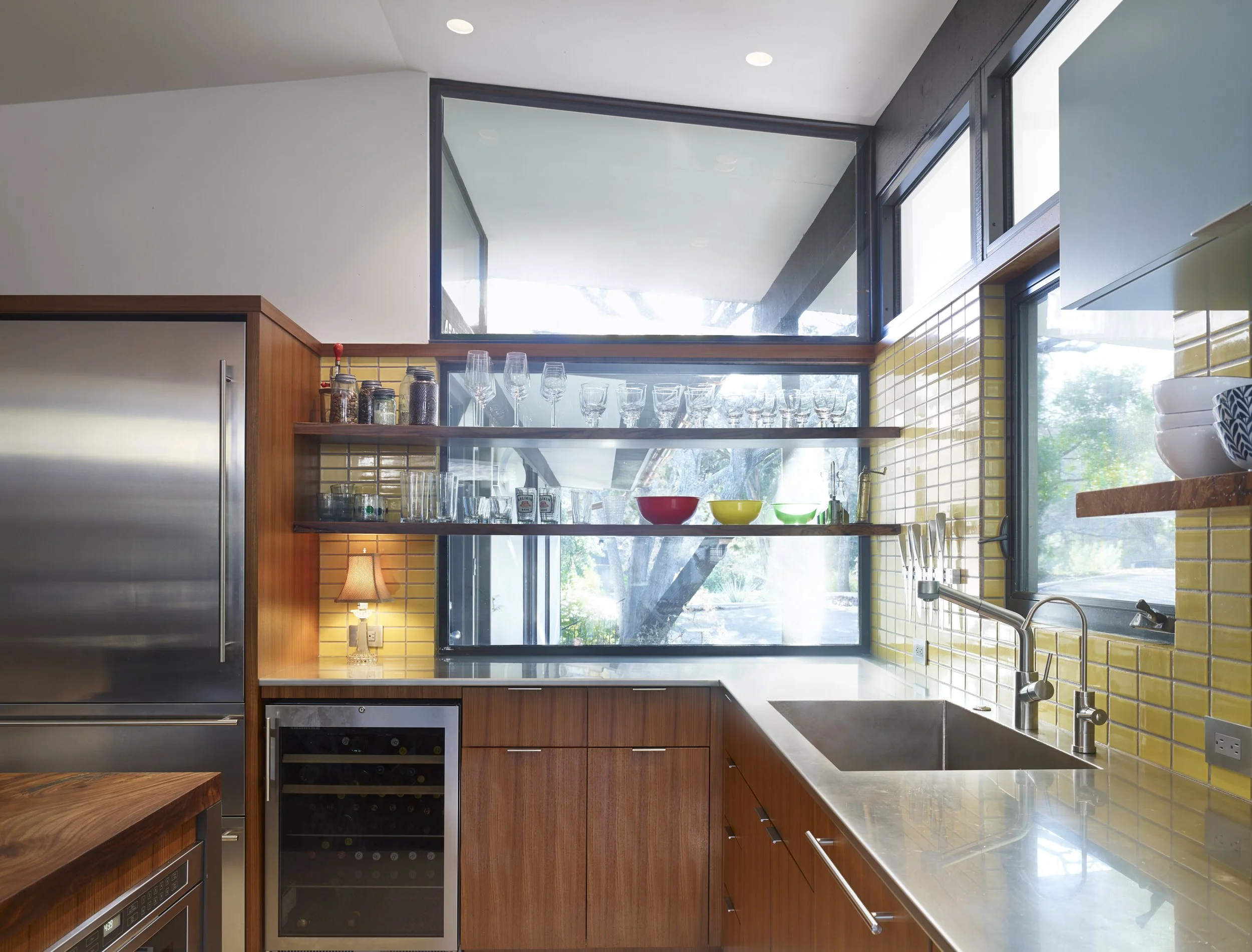 Austin architecture. Modern kitchen with yellow tile backsplash, wooden cabinets, open glass shelves with glassware and bowls, large window, and stainless steel appliances.