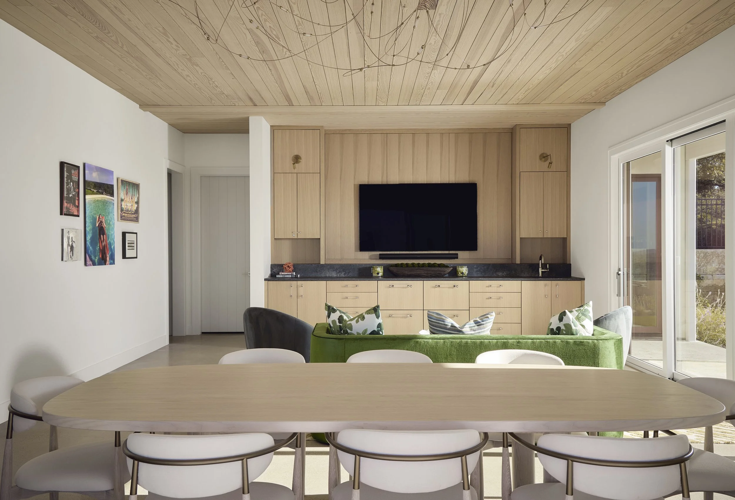 Living room with a light wood ceiling and cabinets, black countertop, wall-mounted TV, sliding glass door, and a dining table with six white chairs. There is a green sofa with floral and striped pillows, and a wall decorated with several colorful art pieces.