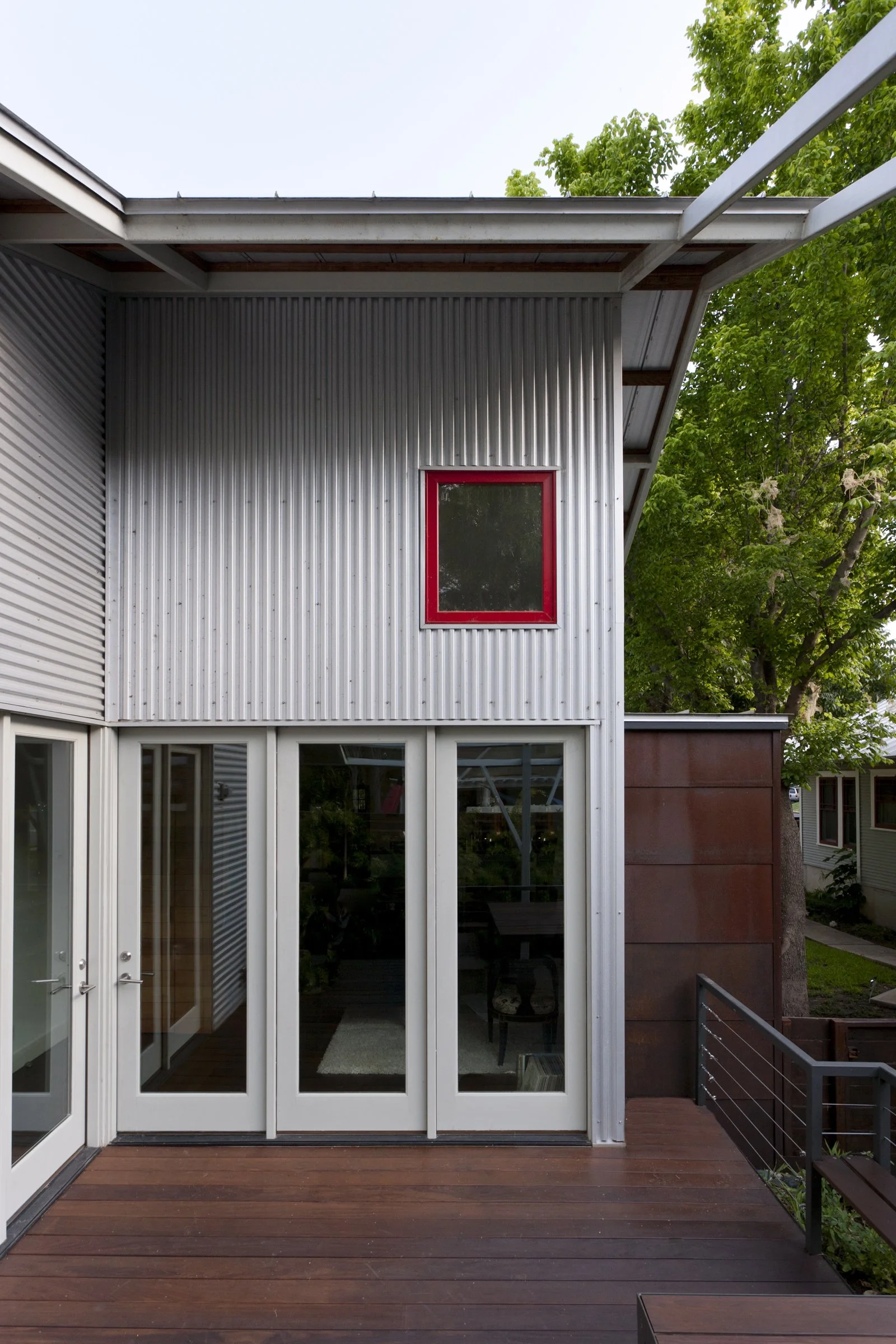 Front of a modern house with metal siding, glass doors, a small red-framed window, a wooden deck, and a large green leafy tree in the background.