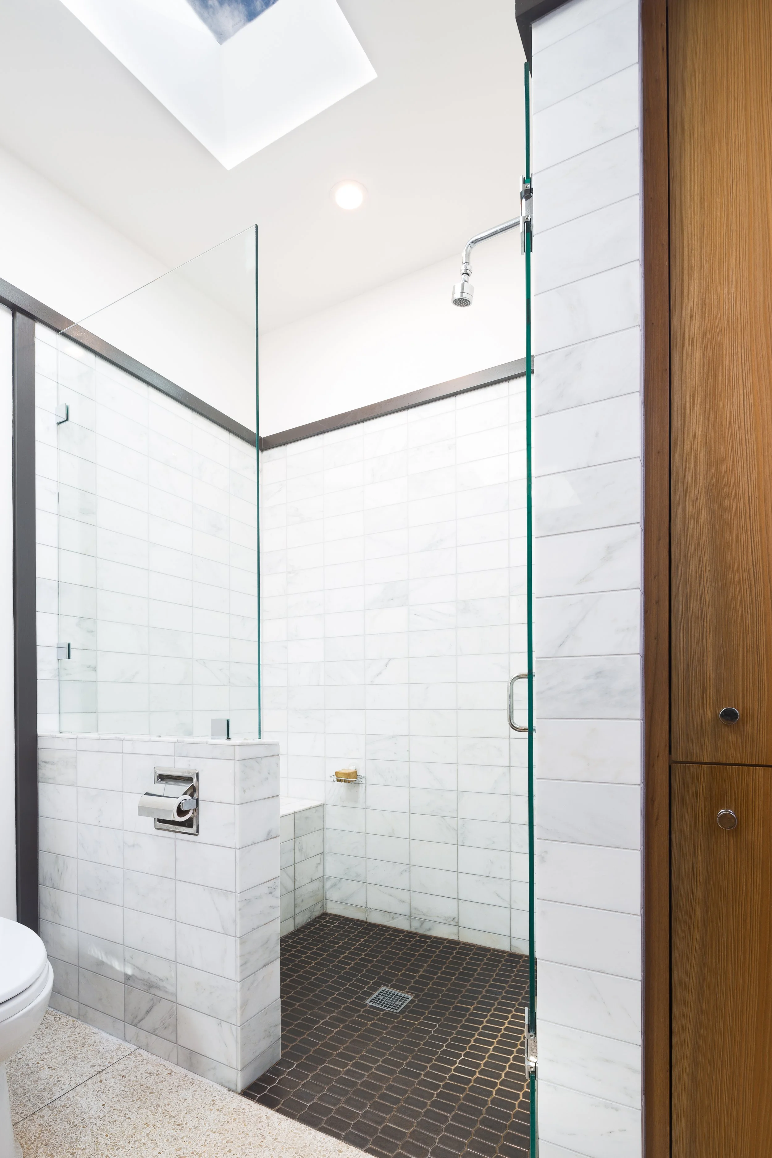 Modern bathroom with white tiled walls, a glass shower enclosure, dark brown mosaic shower floor, and wooden cabinets.