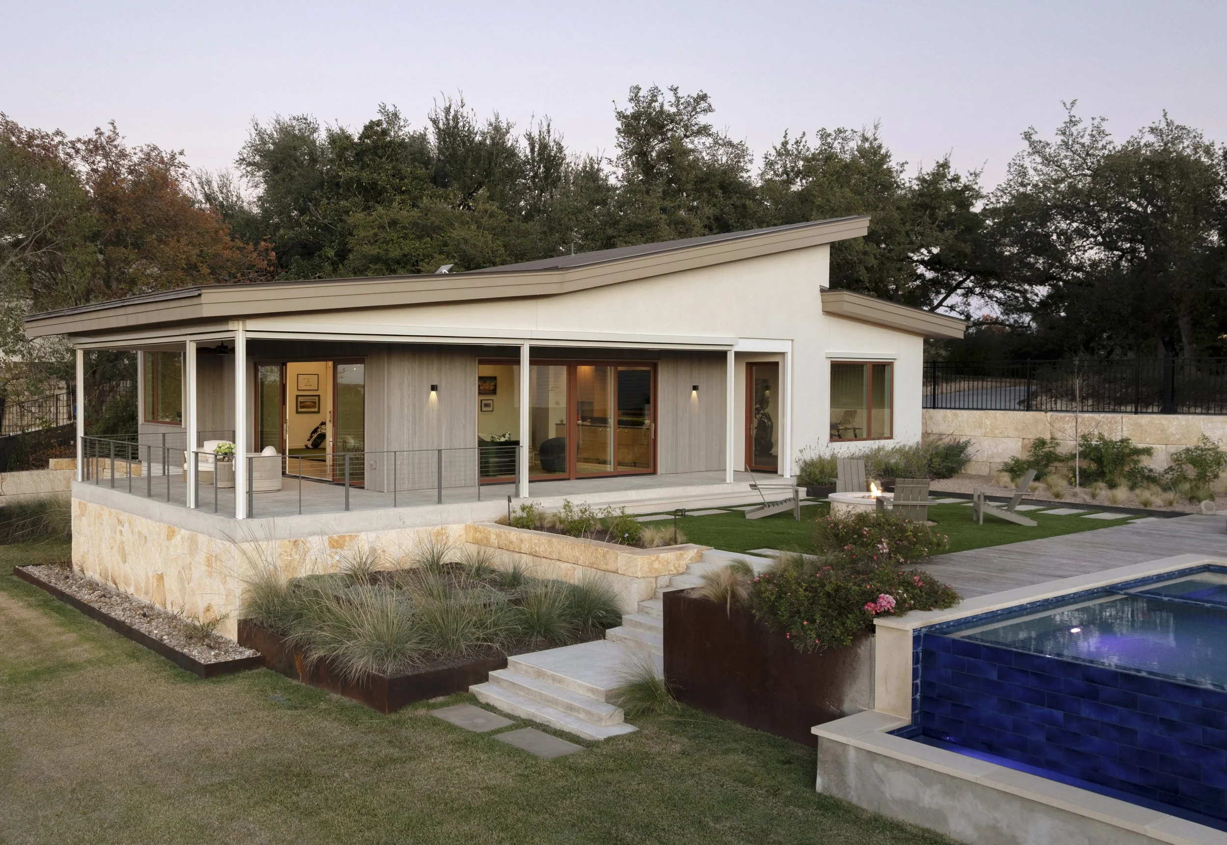 Modern house with a sloped roof, large glass sliding doors, a spacious outdoor patio with seating, and a swimming pool surrounded by a landscaped yard with plants and bushes.