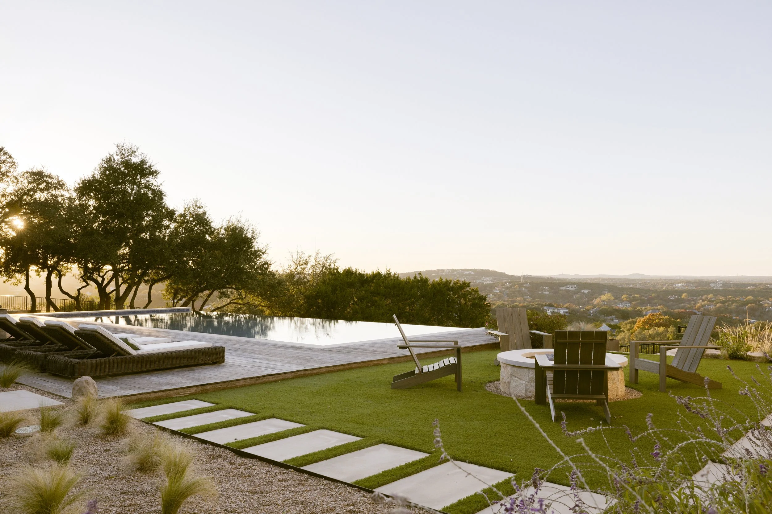 A backyard with a swimming pool, outdoor furniture, a fire pit, and a view of trees and distant landscapes during sunset.