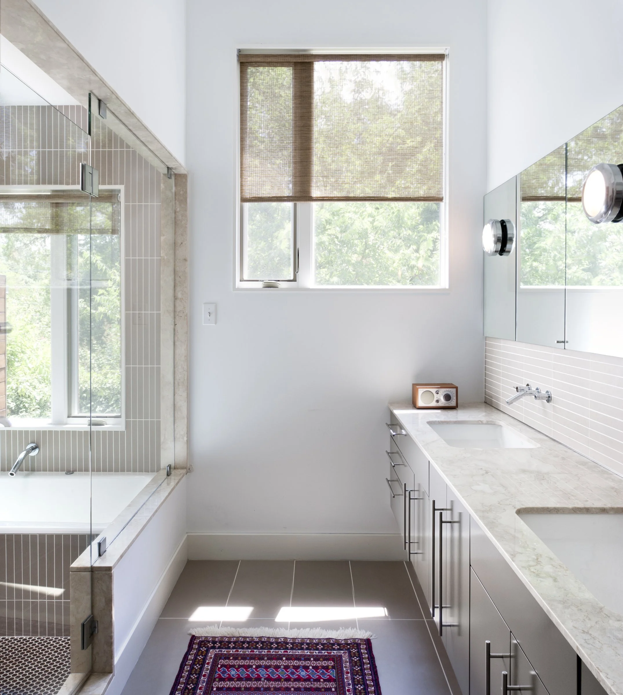 Austin architecture. Modern bathroom with a large window, white cabinets with marble countertops, a small radio on the counter, a wall mirror, and a glass-enclosed bathtub with beige tile accents.