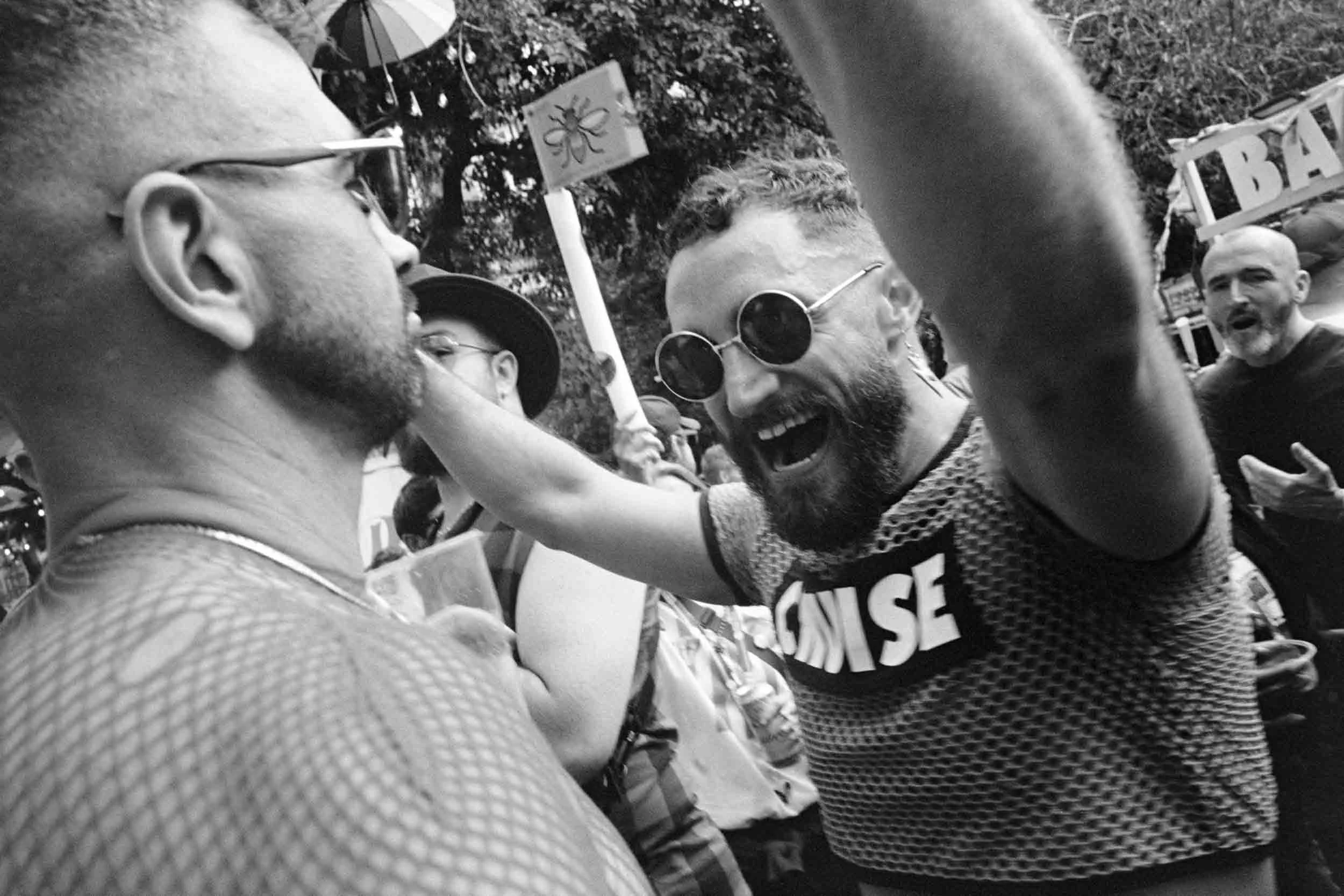 People celebrating outdoors in a protest or rally, one person with a beard and sunglasses smiling and embracing another person wearing a mesh shirt, with signs visible in the background.