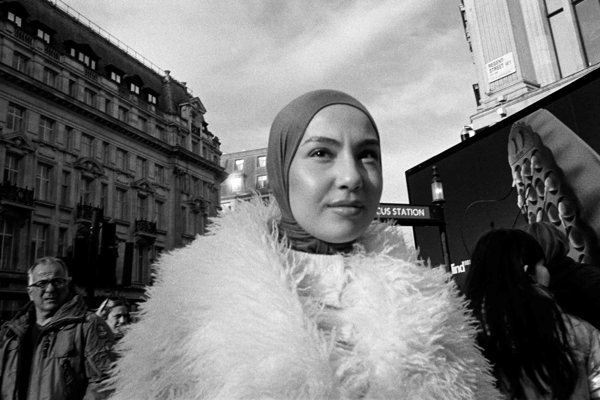 A woman with a hijab standing in an urban street surrounded by tall buildings, along with other people in the background.