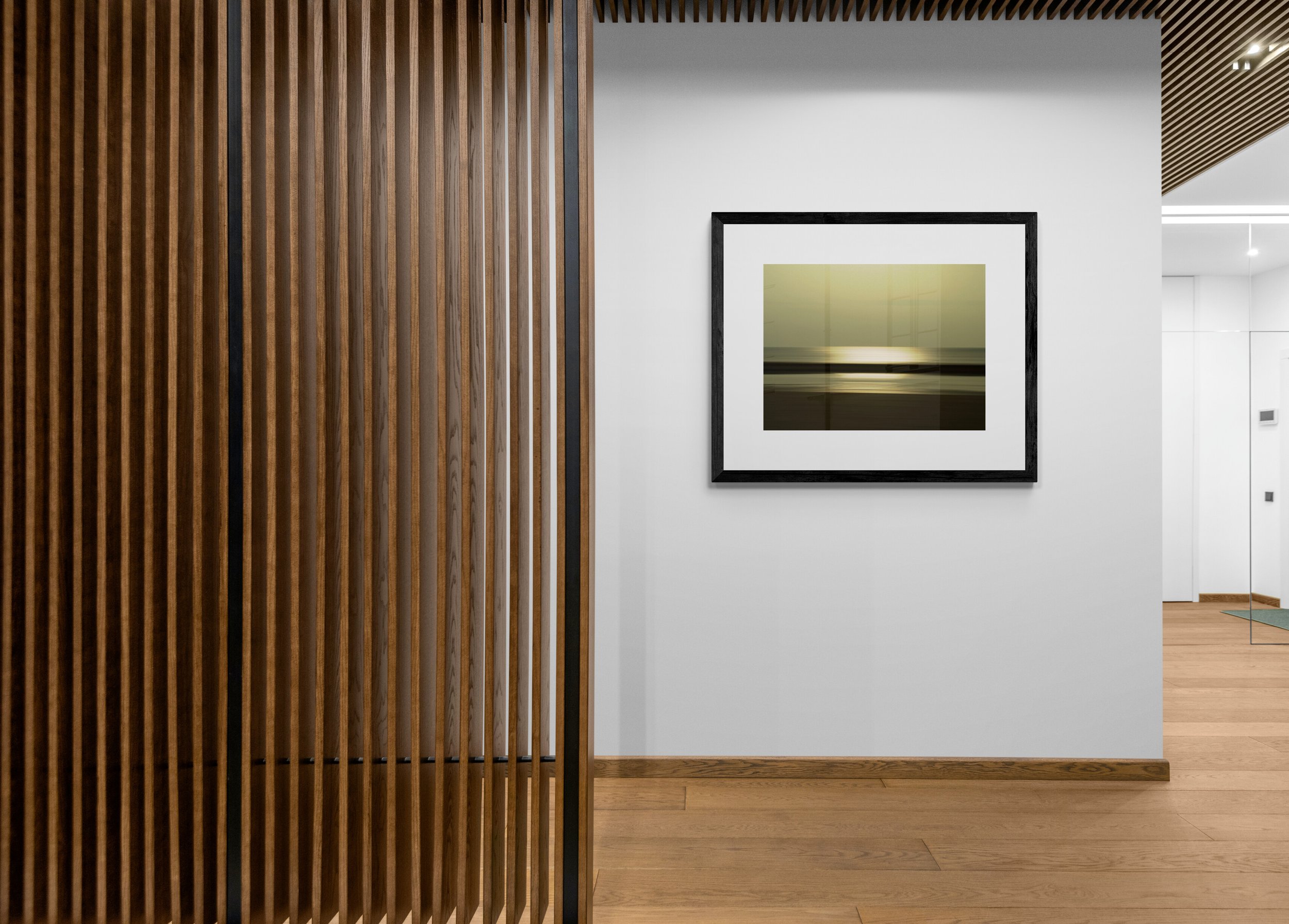 Interior view of an art gallery or modern home, featuring a wooden slat room divider, framed artwork on a white wall, and wooden flooring.