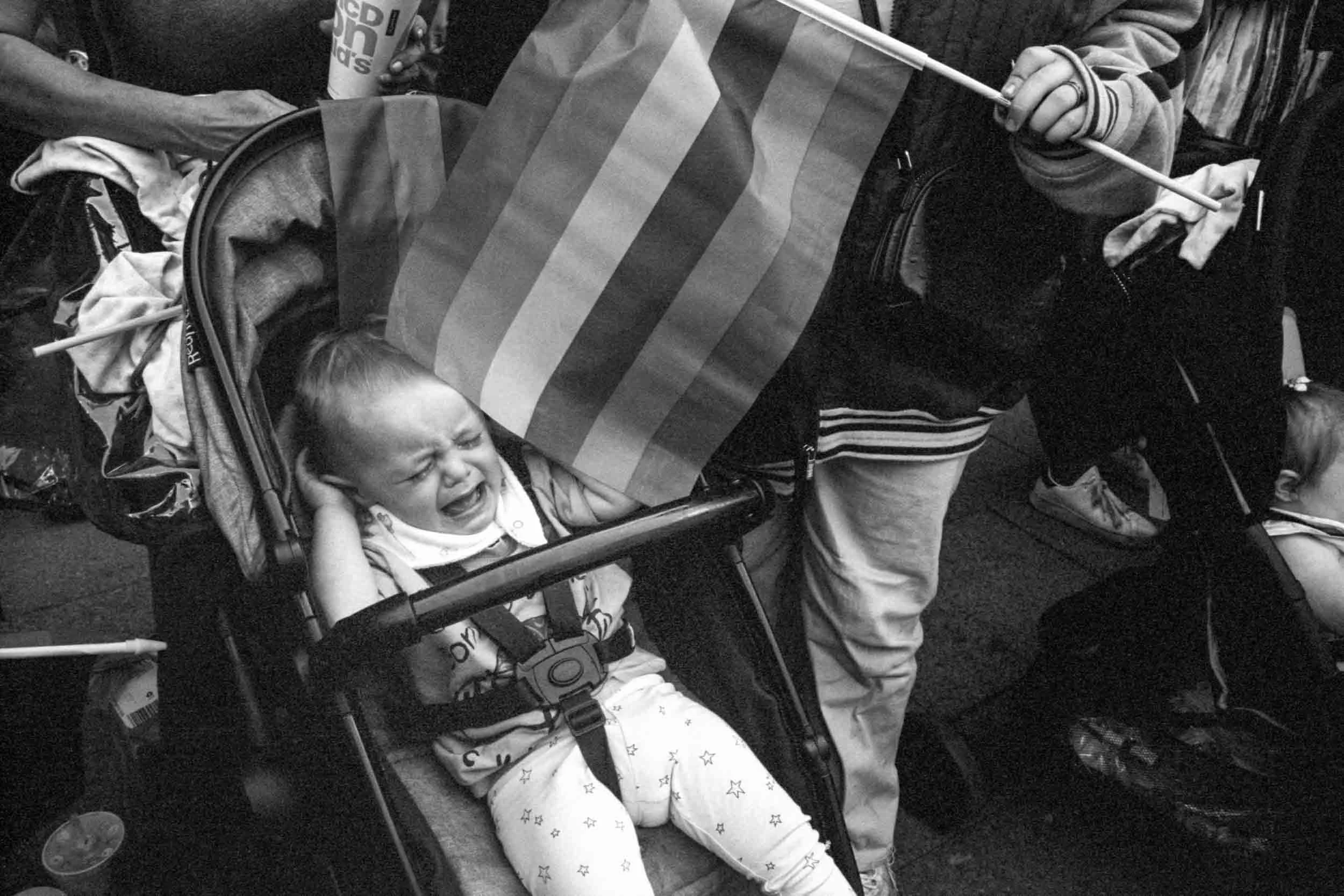 A child sitting in a stroller crying, with a person standing next to them holding a flag and a cup, surrounded by other people.
