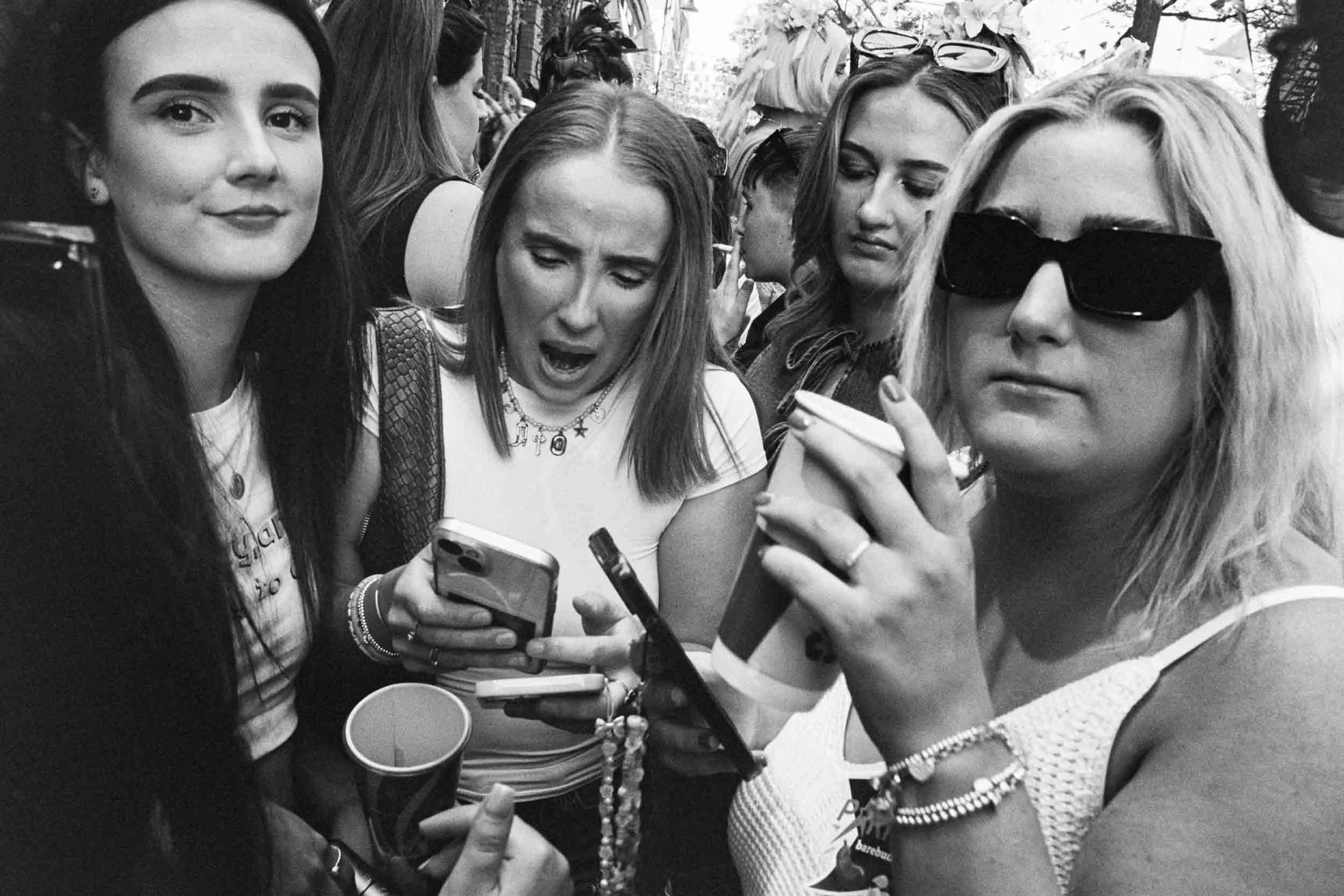 Group of young women outdoors, some wearing sunglasses, engaged with their smartphones, in black and white photo.