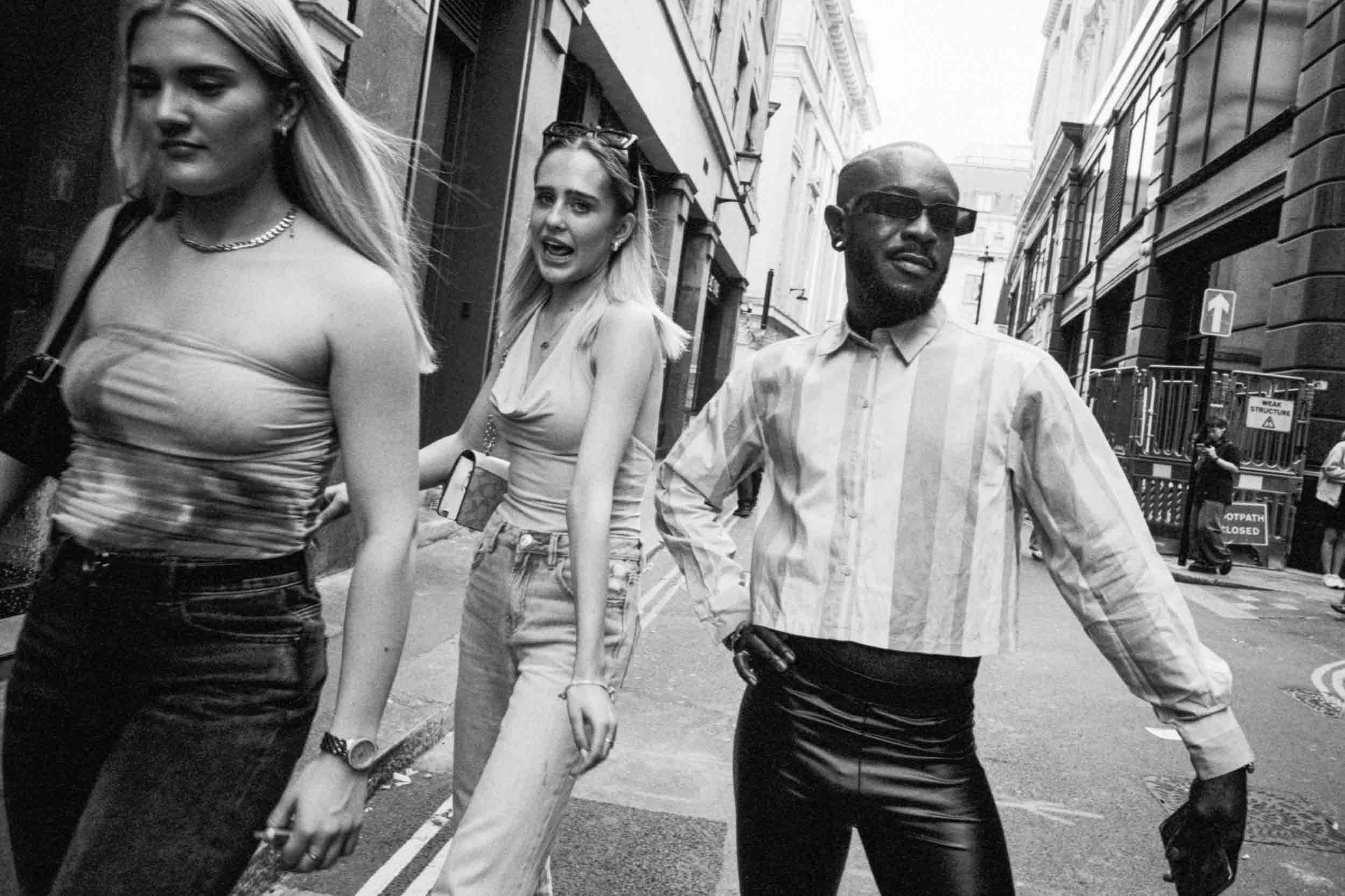 Three young adults, two women and one man, walking down a city street holding hands. The woman on the left has long blonde hair, wearing a strapless top and denim skirt, carrying a purse. The woman in the middle has her hair up, wearing a sleeveless 