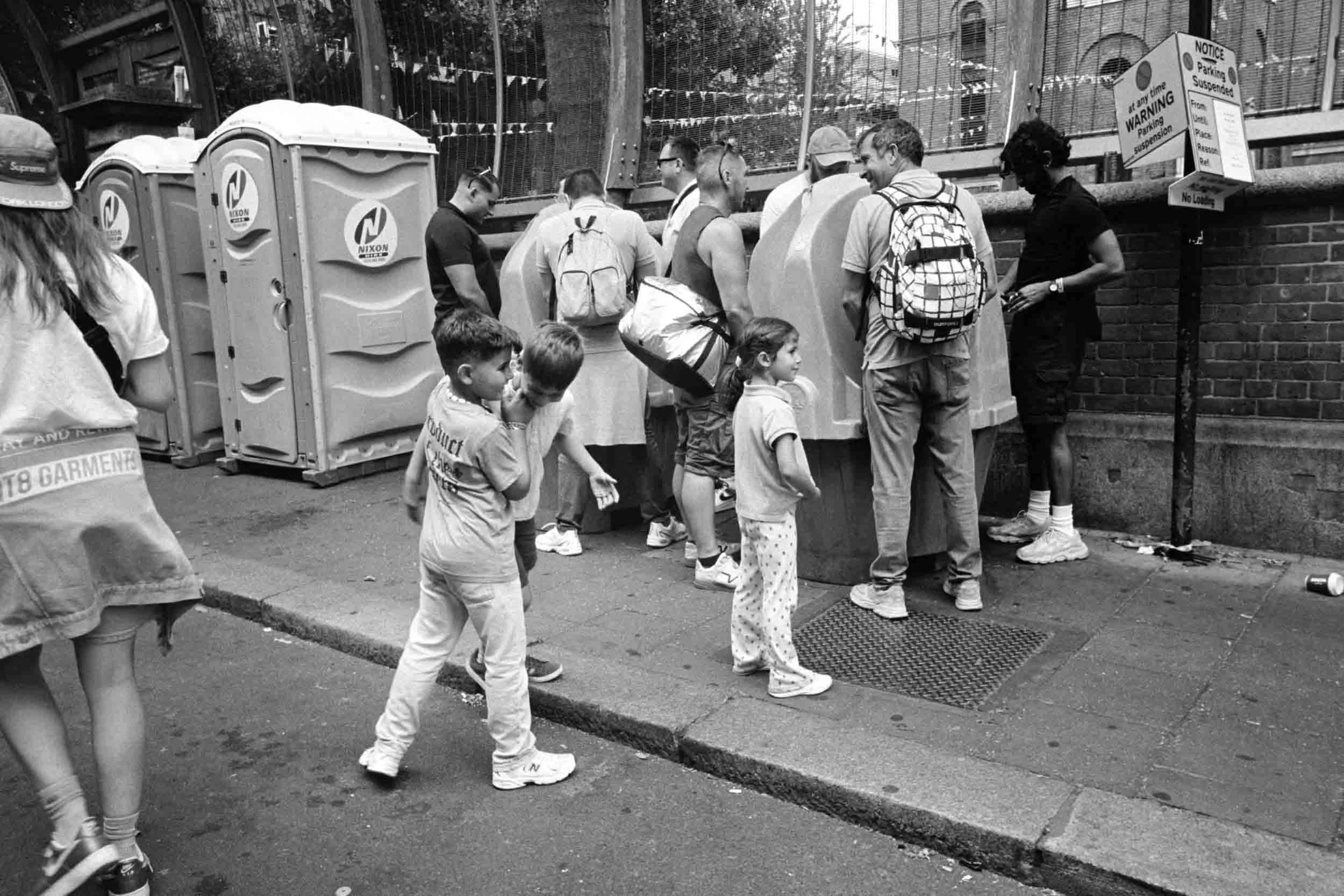A group of people standing in line on the sidewalk outside a building, with some children and adults, near portable toilets and a sign indicating parking restrictions.