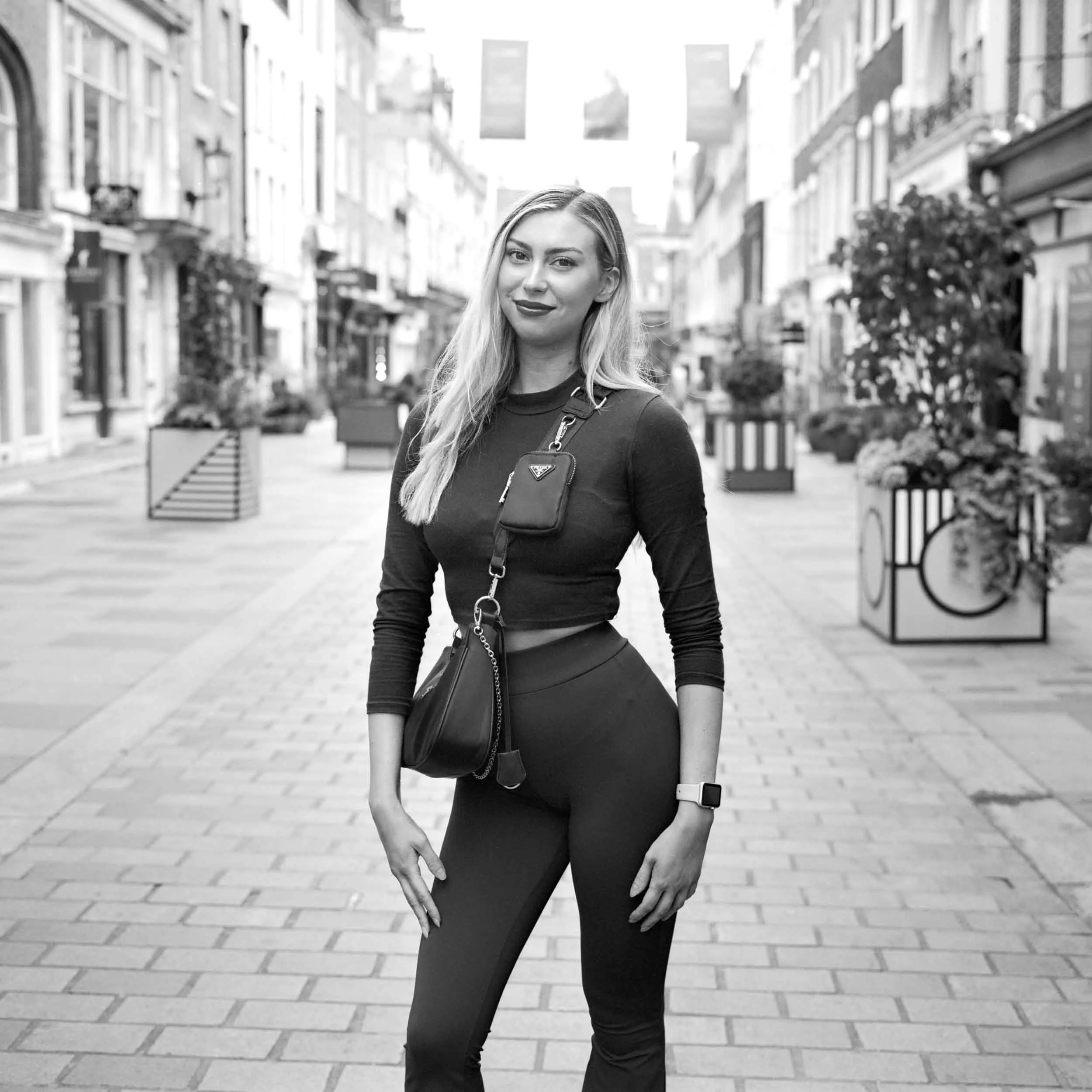 A woman with long blonde hair wearing a black shirt and tights standing on a city sidewalk, holding a small Prada purse, with urban buildings and decorative planters in the background.