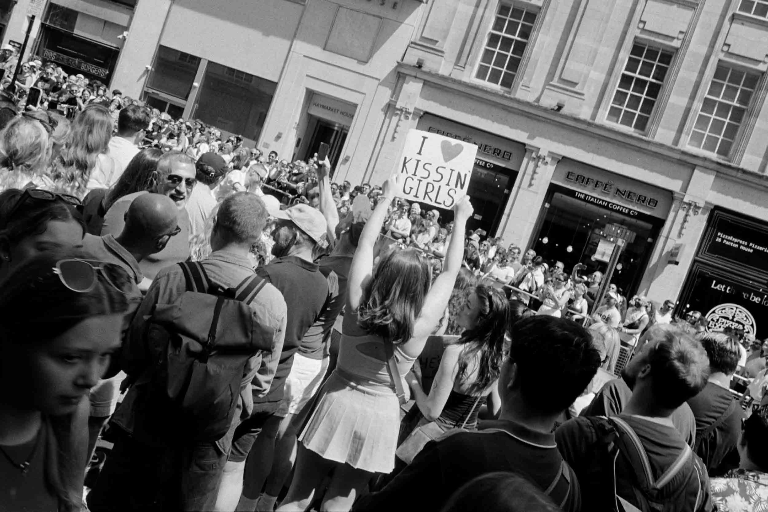 A crowd of people gathered outdoors on a city street, with some holding signs including one that reads 'I ♥ KISSING GIRLS', in front of buildings with large windows and a cafe.